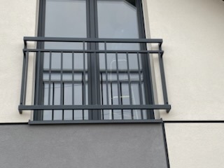 Szara, metalowa balustrada okienna z prostych profili, zamontowana na elewacji budynku z oknem w ciemnej ramie. Minimalistyczny design, nowoczesne budownictwo.