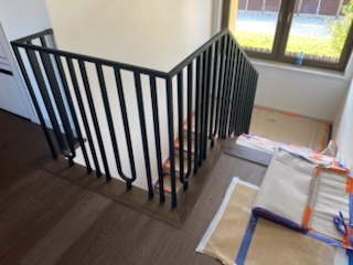 Nowoczesne schody z czarną, metalową balustradą o pionowych szczeblach, widoczne z góry, z zabezpieczonymi stopniami i podłogą. Minimalistyczny design.