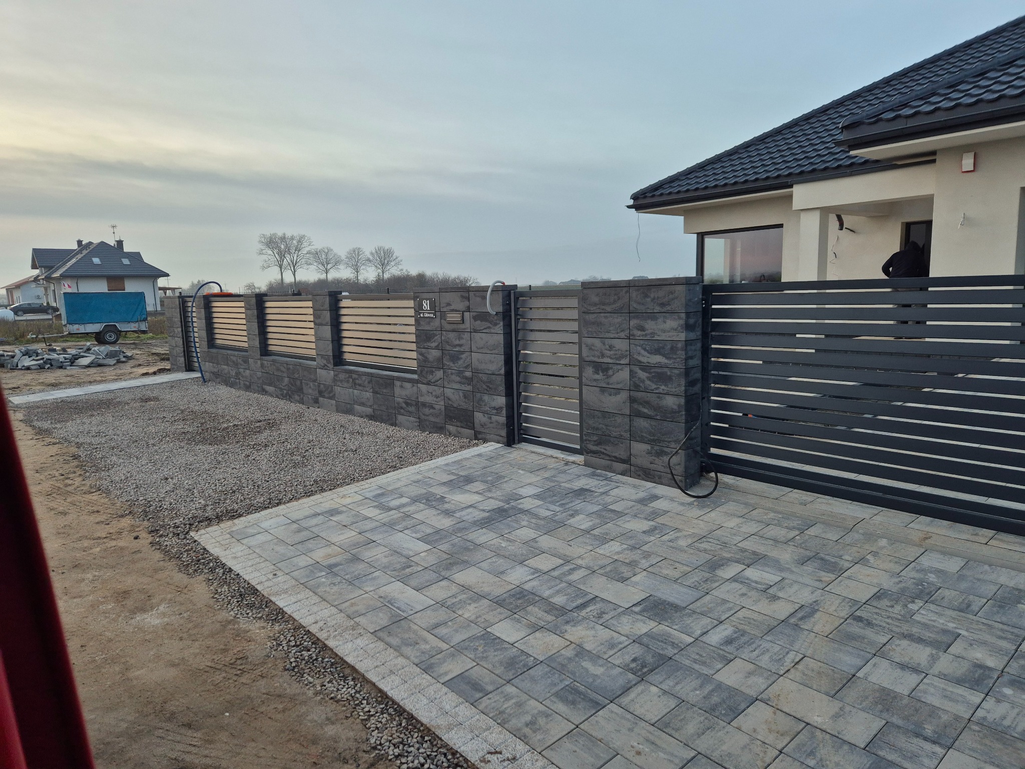 Nowoczesne ogrodzenie posesji z przesuwną bramą wjazdową, wykończone szarą kostką brukową. Widoczny fragment domu z ciemnym dachem i jasną elewacją.