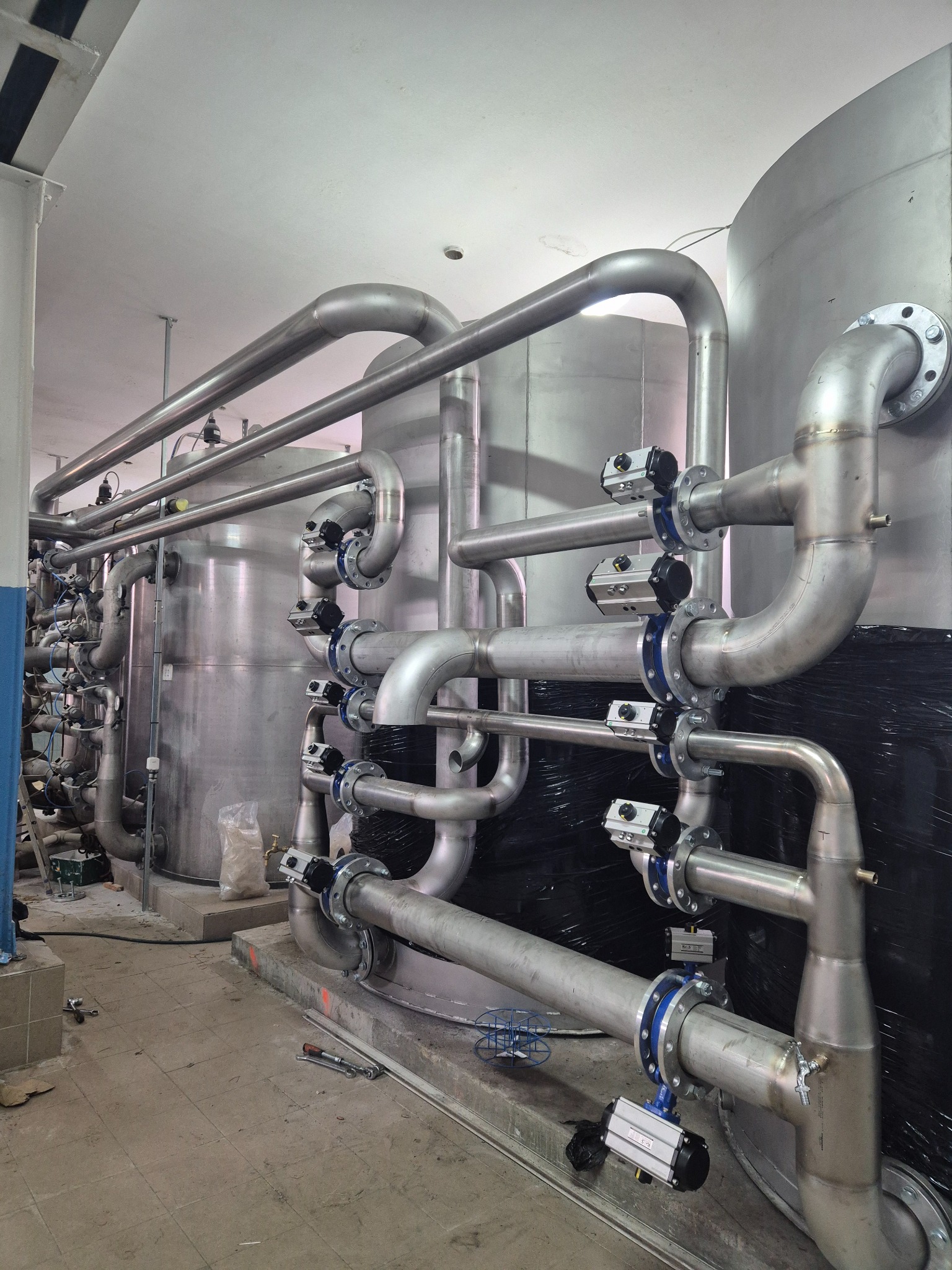 Kompleksowy system rur ze stali nierdzewnej z zaworami, połączonych niebieskimi kołnierzami, w hali przemysłowej. Widoczne zbiorniki i narzędzia.
