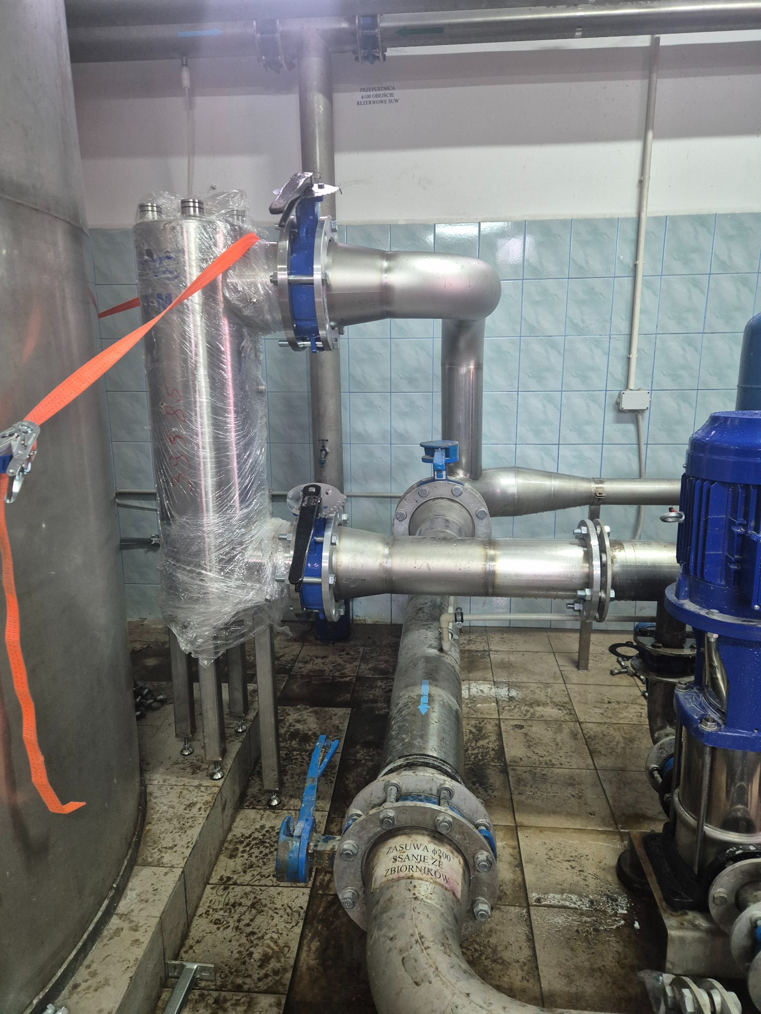 Instalacja przemysłowa z rurami ze stali nierdzewnej, zaworami i zbiornikiem, częściowo owinięta folią ochronną. Widoczne elementy mocujące i silnik pompy.