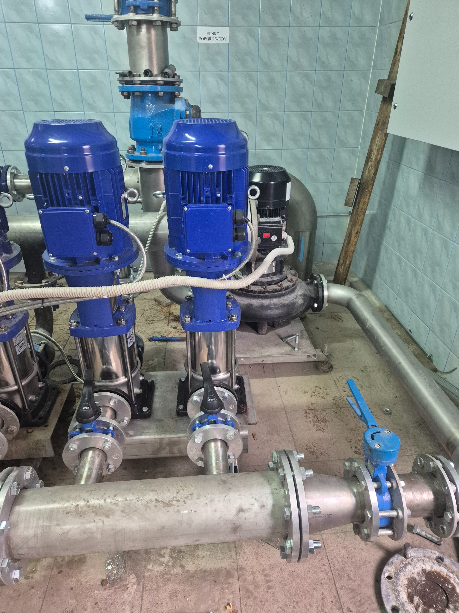 Instalacja pomp i rur w pomieszczeniu technicznym w Zambrowie. Widoczne niebieskie pompy i srebrne rury z zaworami. Tło stanowią kafelki na ścianie.