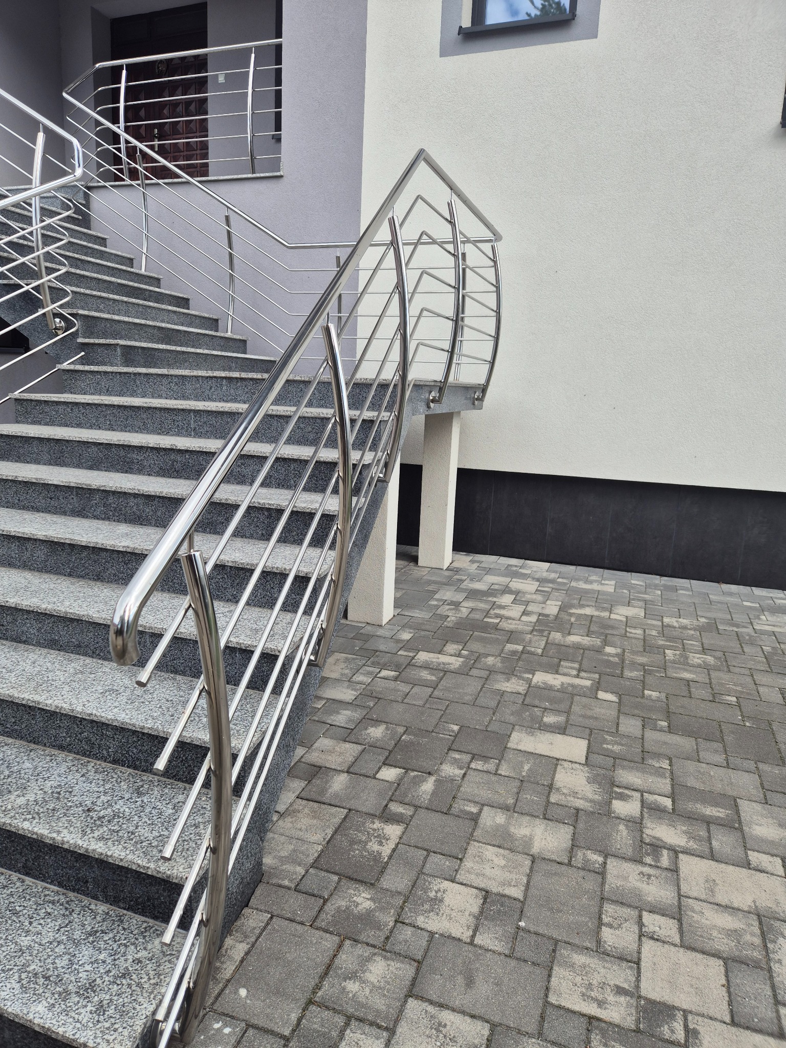 Zewnętrzne schody z szarego granitu z nowoczesną, błyszczącą balustradą ze stali nierdzewnej. Widok z dołu, na pierwszym planie kostka brukowa.