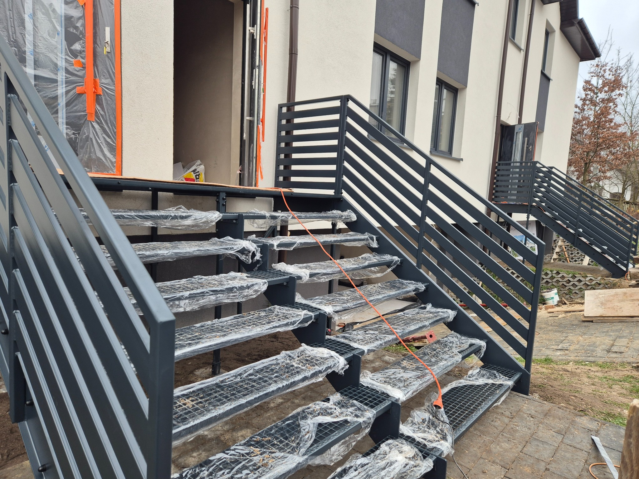 Nowoczesne, metalowe schody zewnętrzne z ażurowymi stopniami, zabezpieczone folią ochronną, prowadzące do wejścia budynku. Minimalistyczne balustrady z poziomych profili.