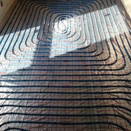 Hydro-Instal Usługi Hydrauliczne Kamil Smolak - Niebieskie rury ogrzewania podłogowego rozłożone spiralnie na folii z czerwoną kratą. Widok z góry, instalacja przed wylaniem posadzki.