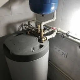 Zamontowany bojler z niebieskim zbiornikiem wyrównawczym i instalacją z rur PCV w pomieszczeniu z białymi płytkami. Widoczne gniazdka elektryczne.