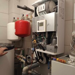Hydro-Instal Usługi Hydrauliczne Kamil Smolak - Biały piec CO Toshiba zamontowany na ścianie z szarą glazurą, widoczne rury, zbiorniki wyrównawcze oraz miernik elektryczny podczas pracy instalatora.