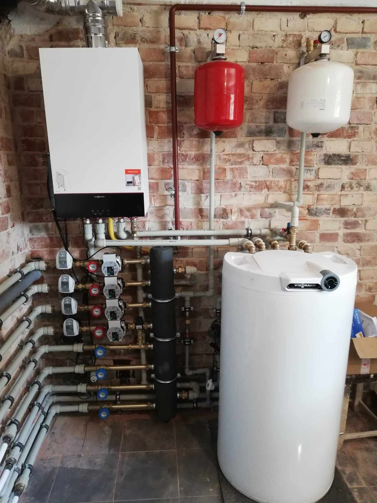 Złożona instalacja grzewcza w pomieszczeniu z ceglaną ścianą: piec gazowy Viessmann, biały zbiornik, czerwony i biały zbiornik wyrównawczy oraz liczne rury i zawory.