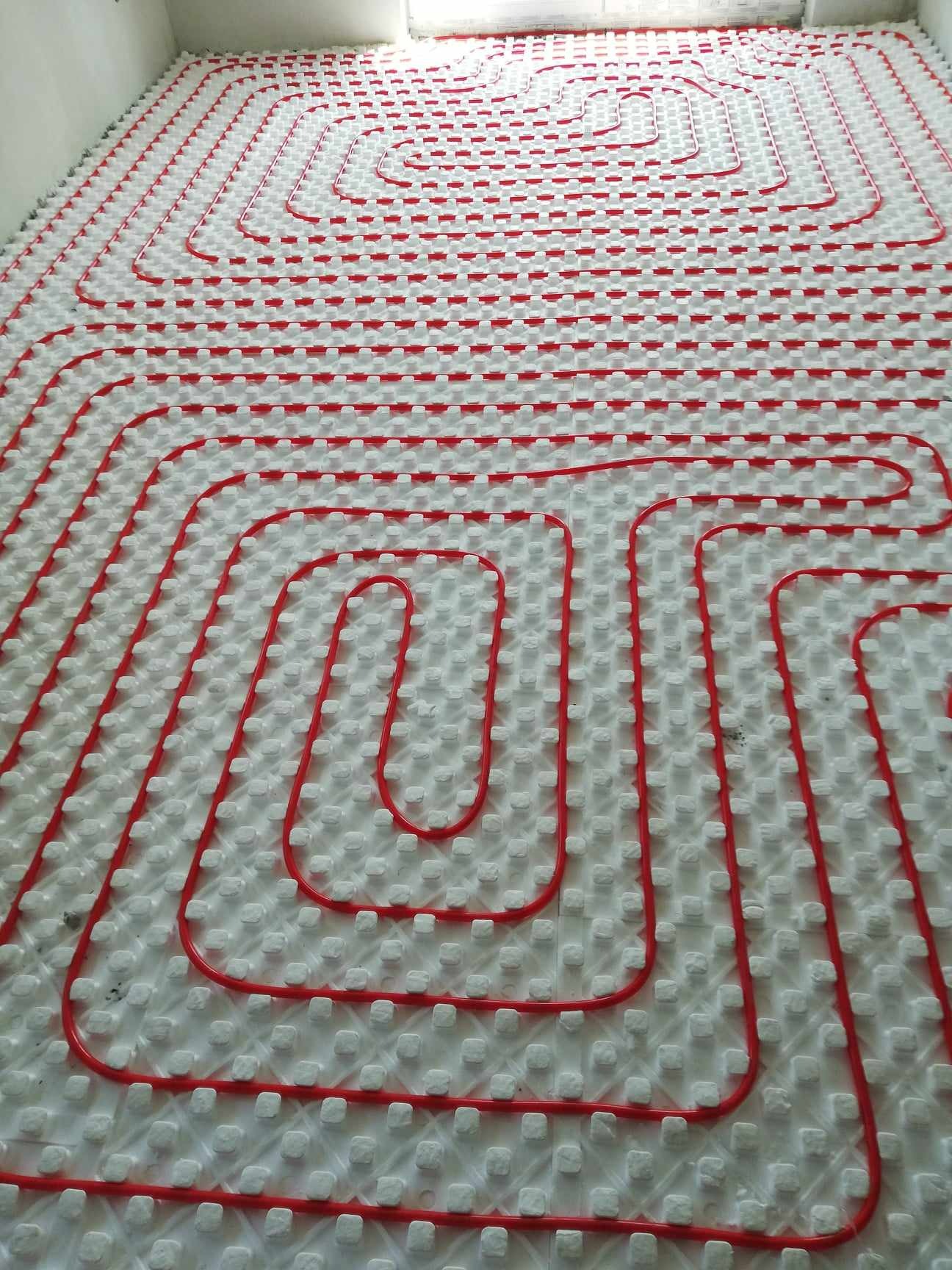 Czerwone rury ogrzewania podłogowego ułożone na białej izolacji z wypustkami, tworzące regularny wzór meandrujący na dużej powierzchni podłogi w jasnym pomieszczeniu.