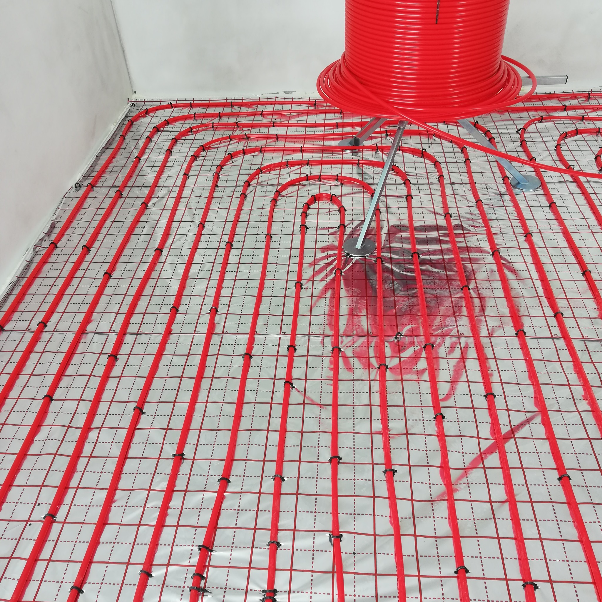 Czerwone rury ogrzewania podłogowego ułożone na folii z rastrem, widoczna rolka rur i narzędzie do ich rozwijania. Instalacja przed zalaniem wylewką.