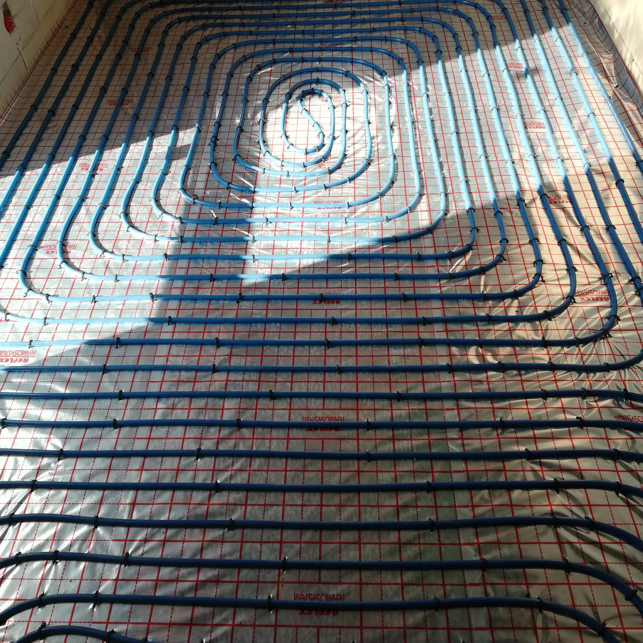 Niebieskie rury ogrzewania podłogowego rozłożone spiralnie na folii z czerwoną kratą. Widok z góry, instalacja przed wylaniem posadzki.