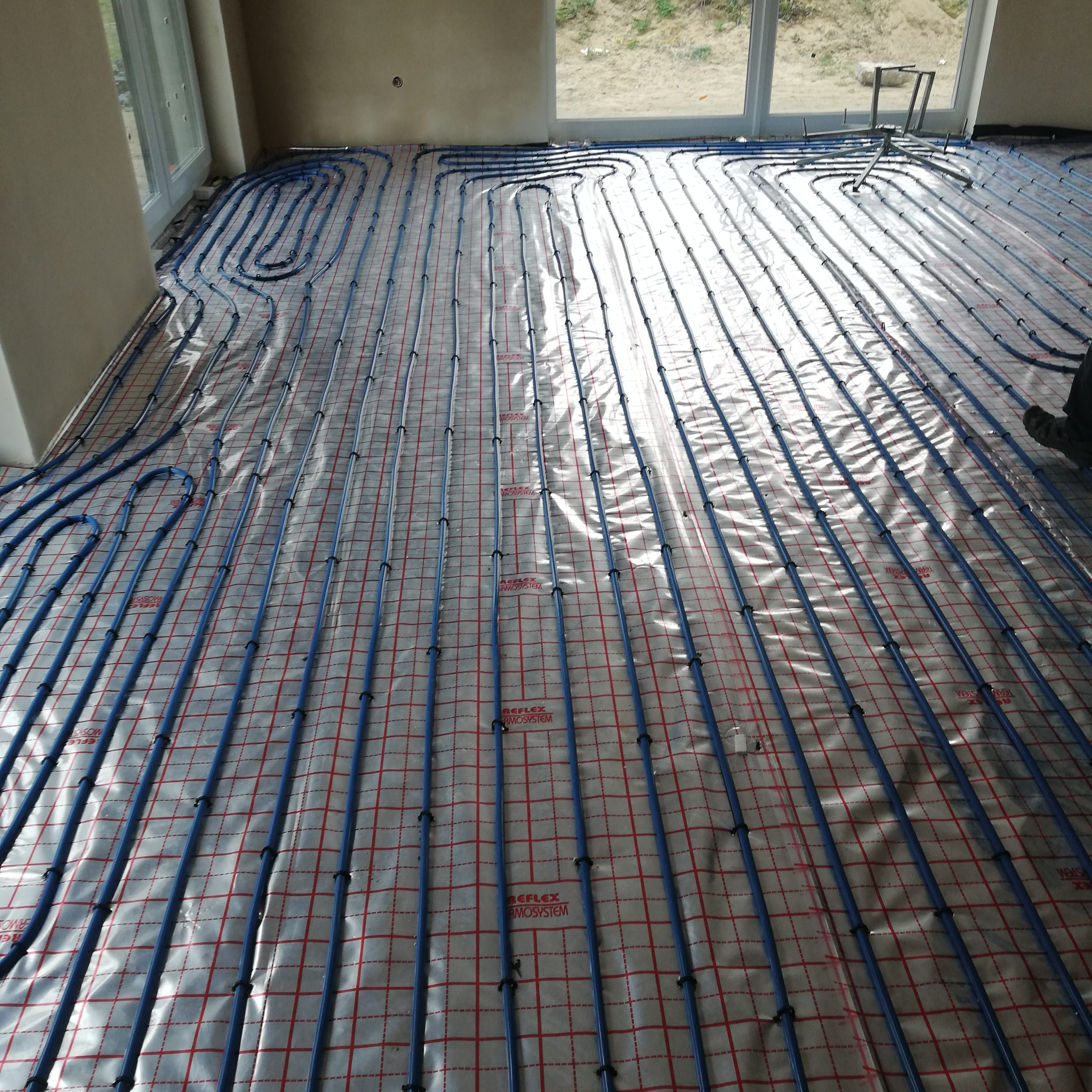 Błękitne rury ogrzewania podłogowego ułożone na folii z czerwoną siatką w jasnym pomieszczeniu, widoczne okna i fragment nogi w rogu.