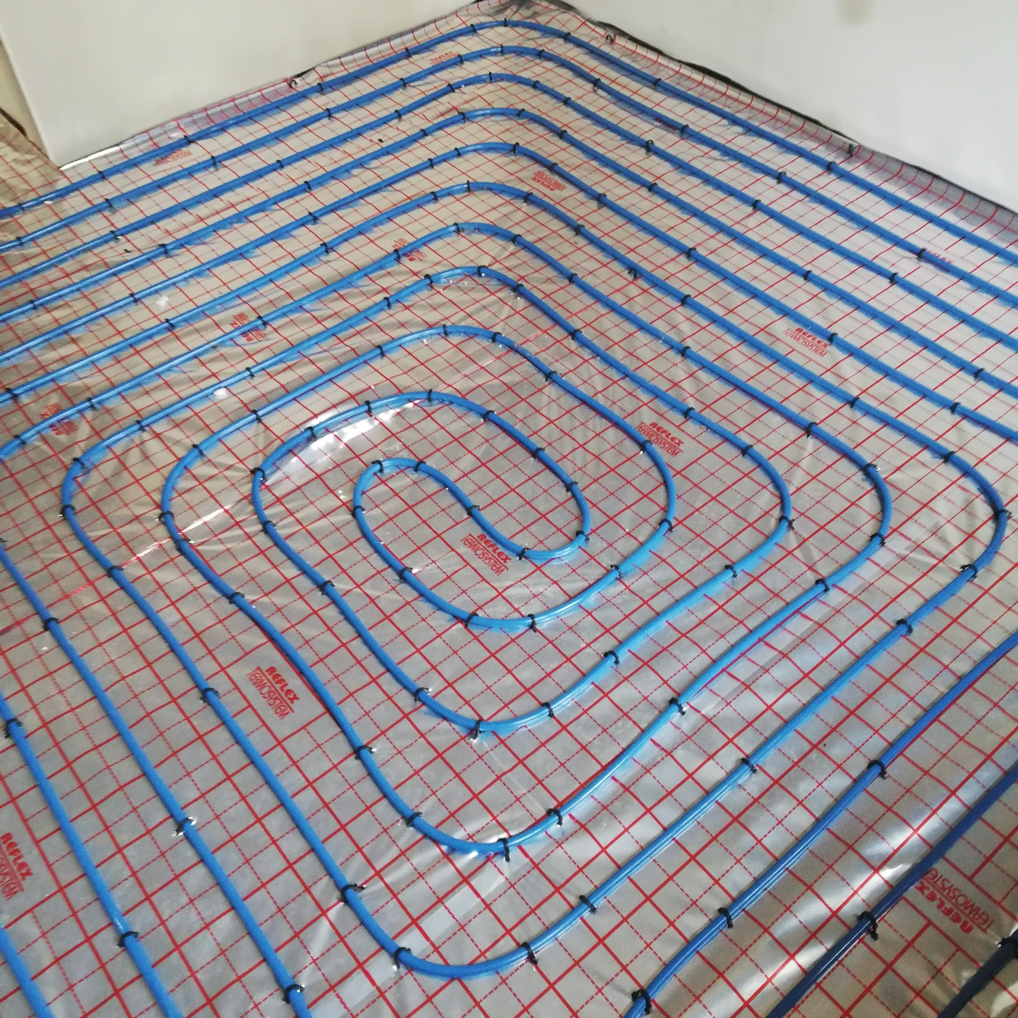 Niebieskie rury ogrzewania podłogowego ułożone w pętle na folii z czerwoną kratką, przymocowane czarnymi uchwytami. Widok z góry na instalację przed zalaniem.