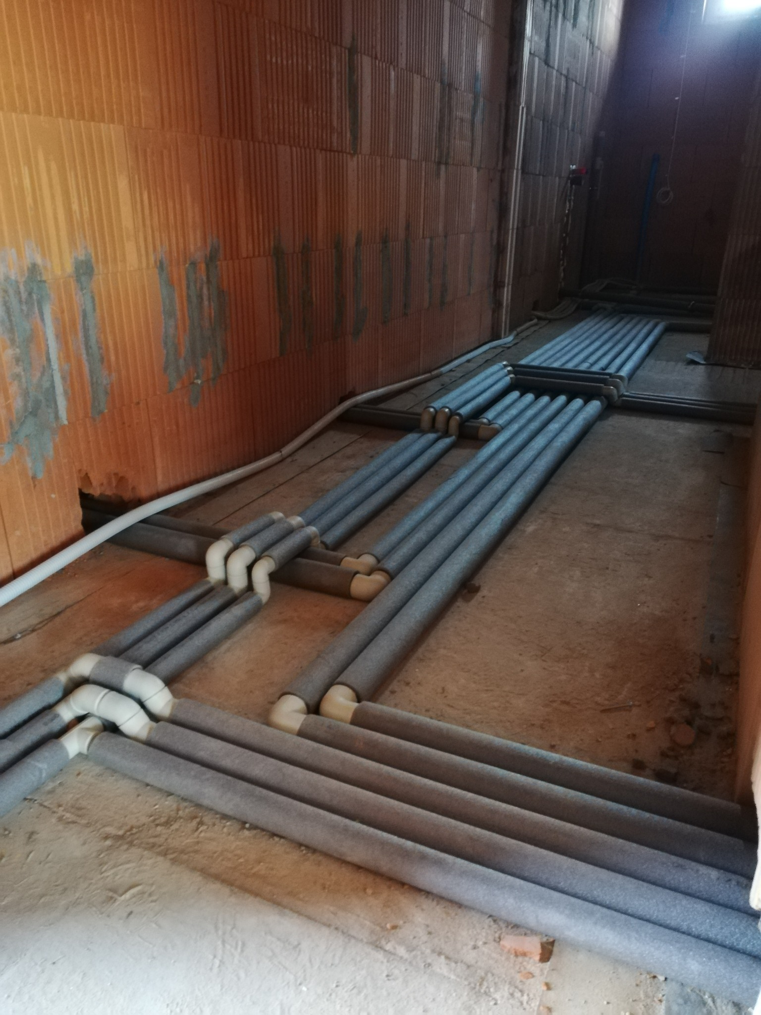 Instalacja rur grzewczych w izolacji, ułożonych na podłożu w surowym wnętrzu budynku z ceglanymi ścianami. Widoczne białe łączniki i fragmenty instalacji na ścianie.