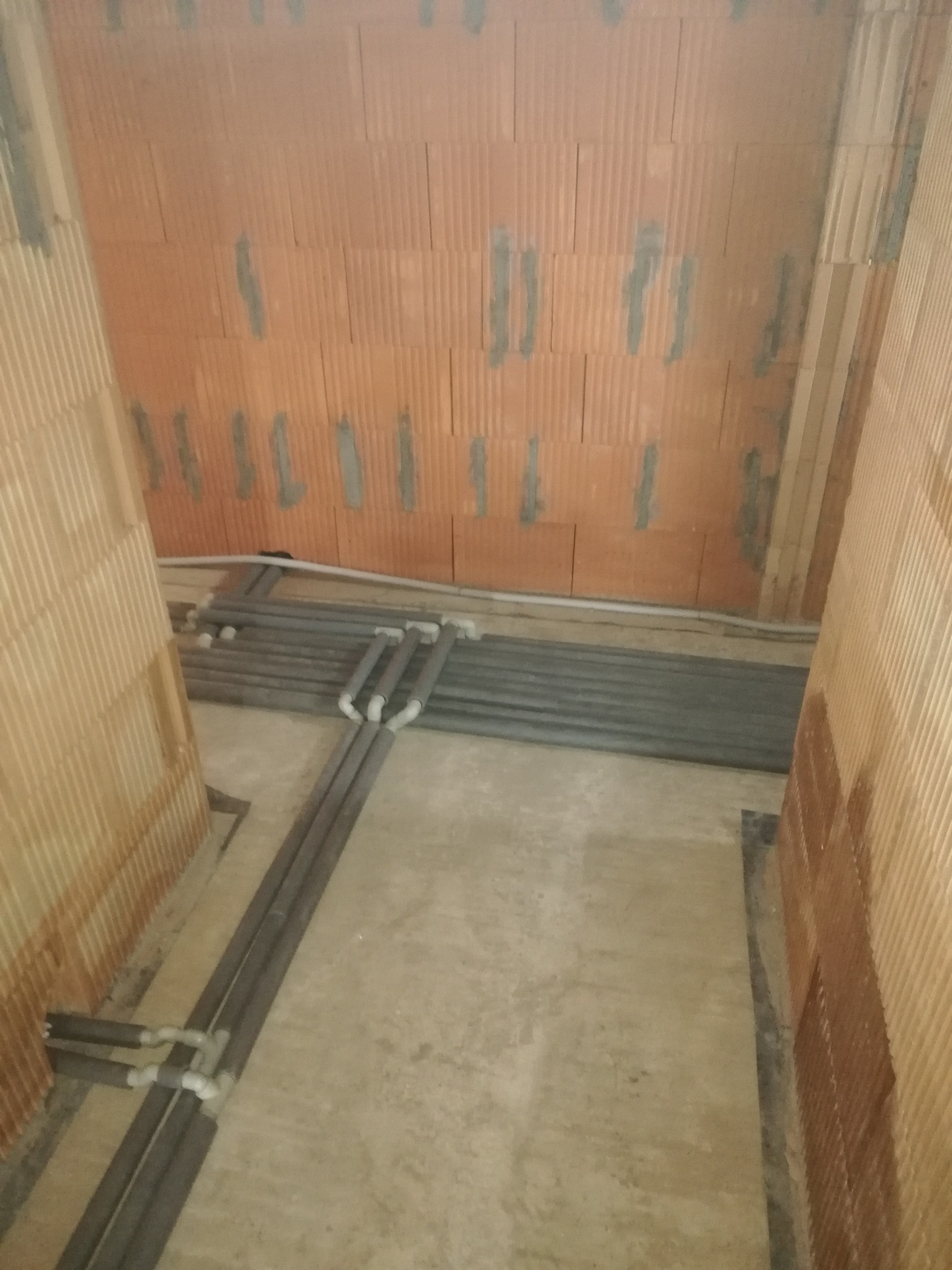 Szare, izolowane rury instalacji grzewczej ułożone na betonowej podłodze w surowym wnętrzu z ceglanymi ścianami. Widok z góry, etap prac przed wylaniem posadzki.