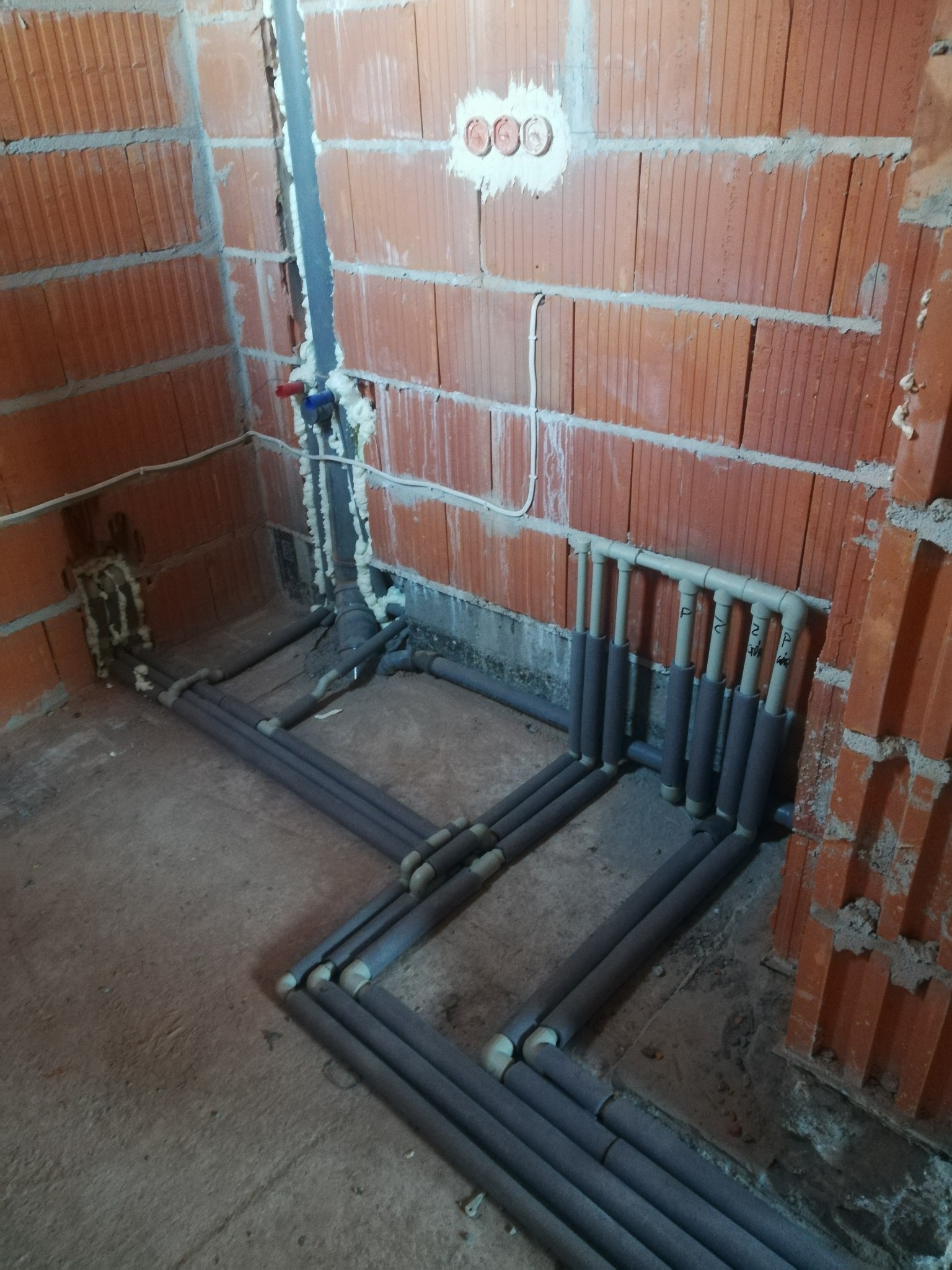 Surowy stan: instalacja ogrzewania podłogowego z szarymi rurami w izolacji termicznej, widoczne ściany z czerwonej cegły i wystające rury pionowe.