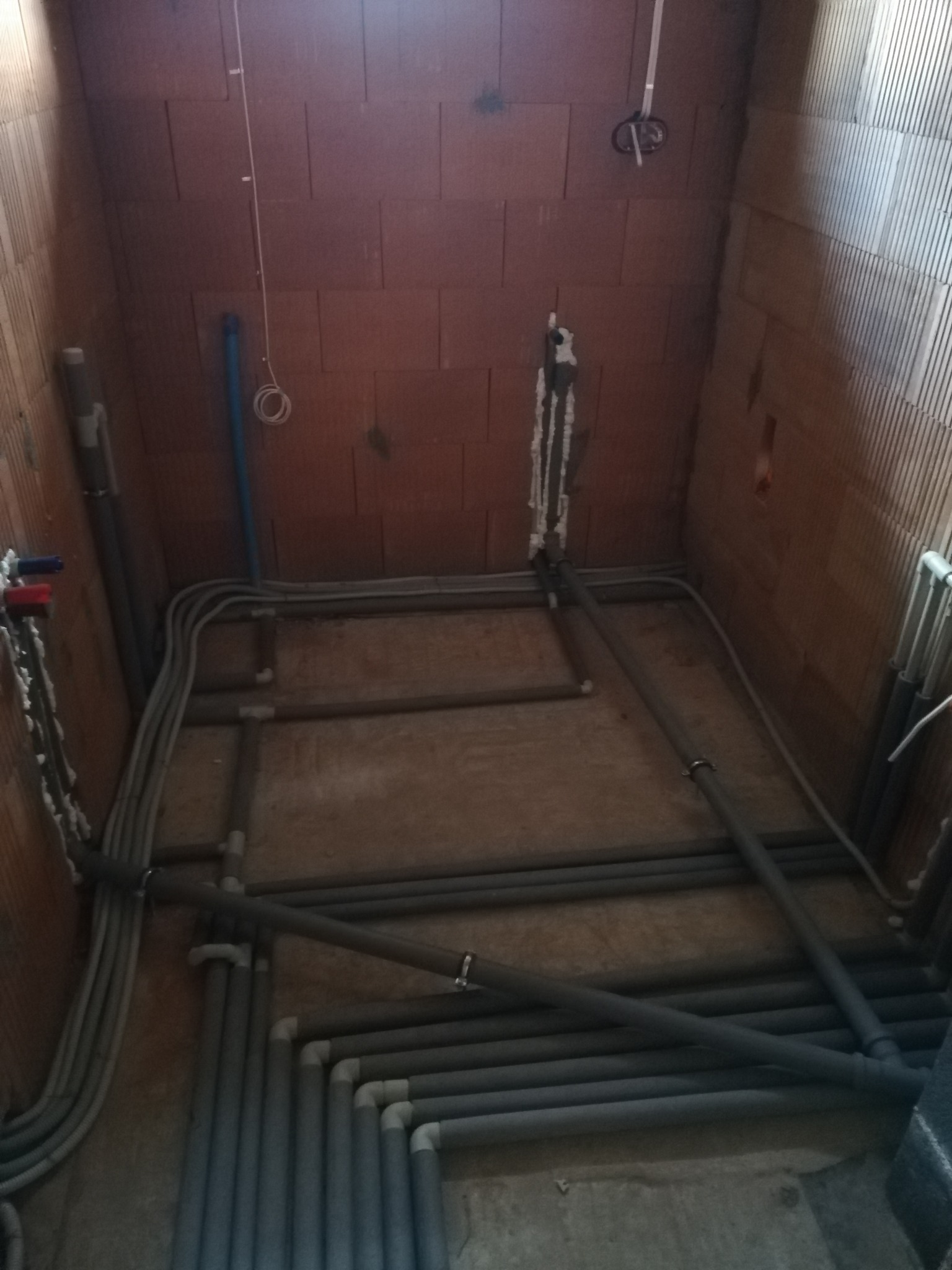 Szare rury instalacji ogrzewania podłogowego ułożone na surowej posadzce w pomieszczeniu z ceglanymi ścianami. Widoczne elementy montażowe i instalacja elektryczna.