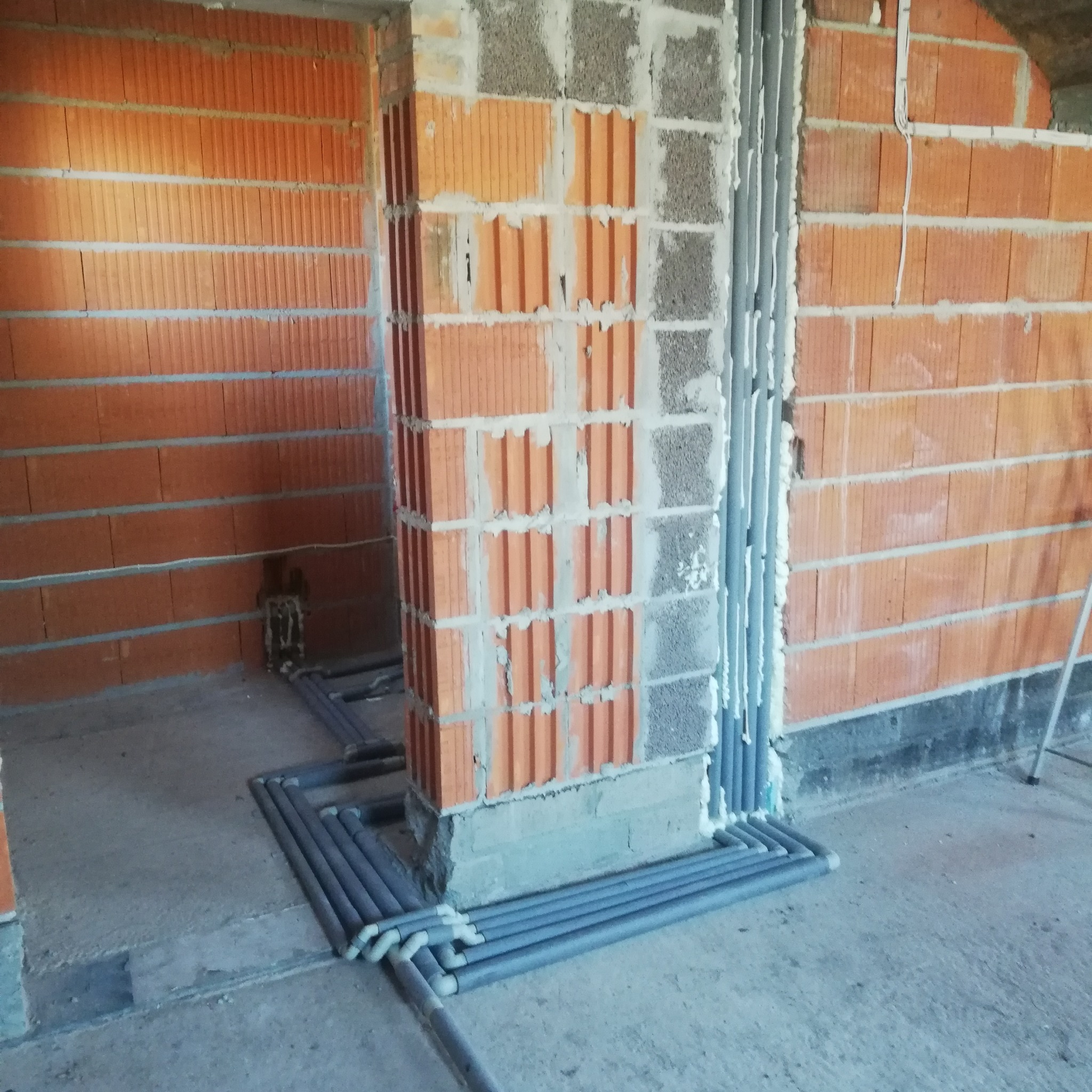 Szare rury instalacji grzewczej ułożone równolegle na podłodze w surowym wnętrzu, widoczne ceglane ściany i fragment instalacji w ścianie.
