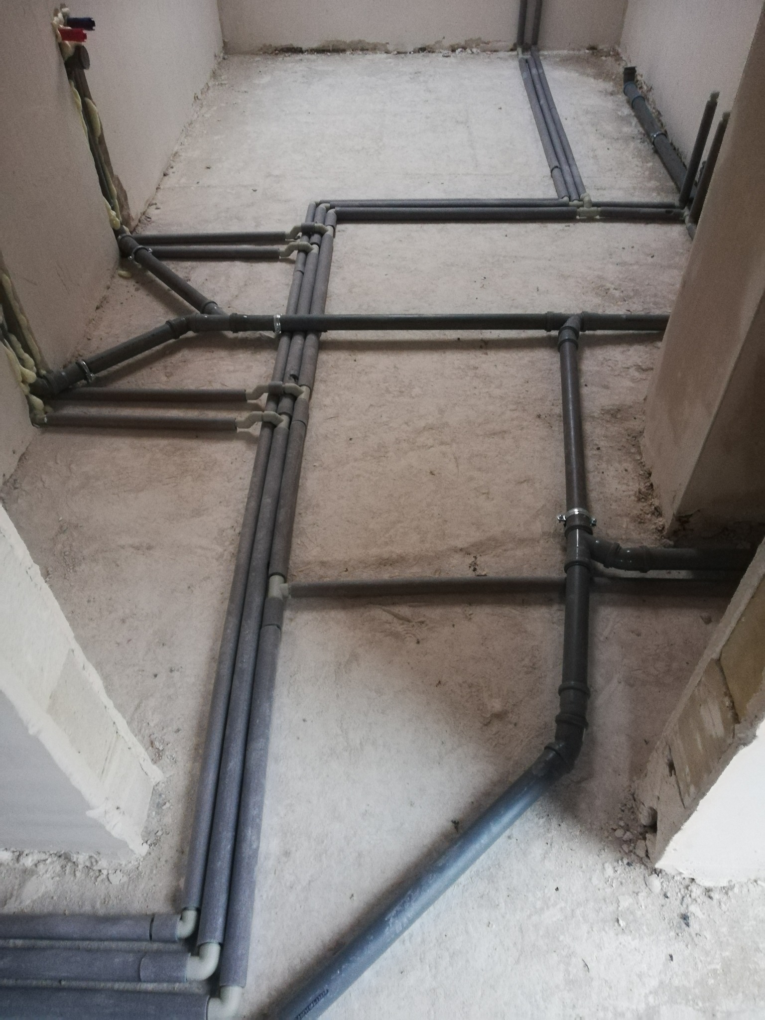 Szare rury instalacji grzewczej ułożone na podłodze w Okuniew. Widoczne łączenia i izolacja termiczna. Świeża wylewka, surowe ściany.