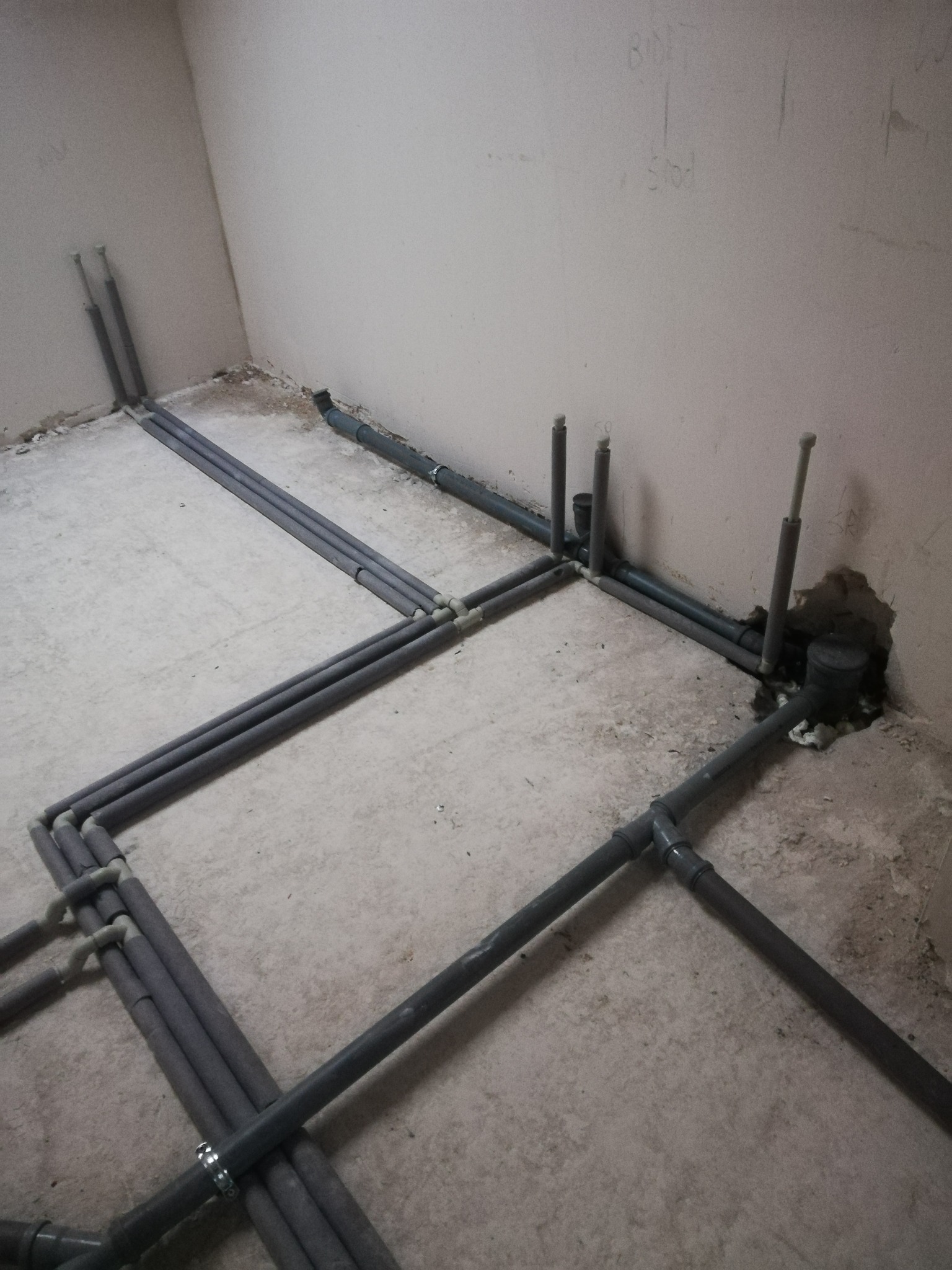 Szare, zaizolowane rury instalacji wodnej ułożone na surowej posadzce w pomieszczeniu z surowymi ścianami. Widoczne pionowe wyprowadzenia rur.