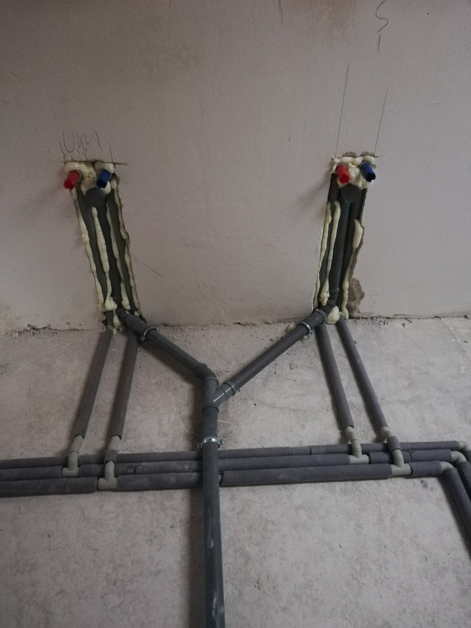 Szare rury instalacji wodnej, zabezpieczone pianką montażową przy ścianie, z czerwonymi i niebieskimi zaślepkami, układane na surowej posadzce.