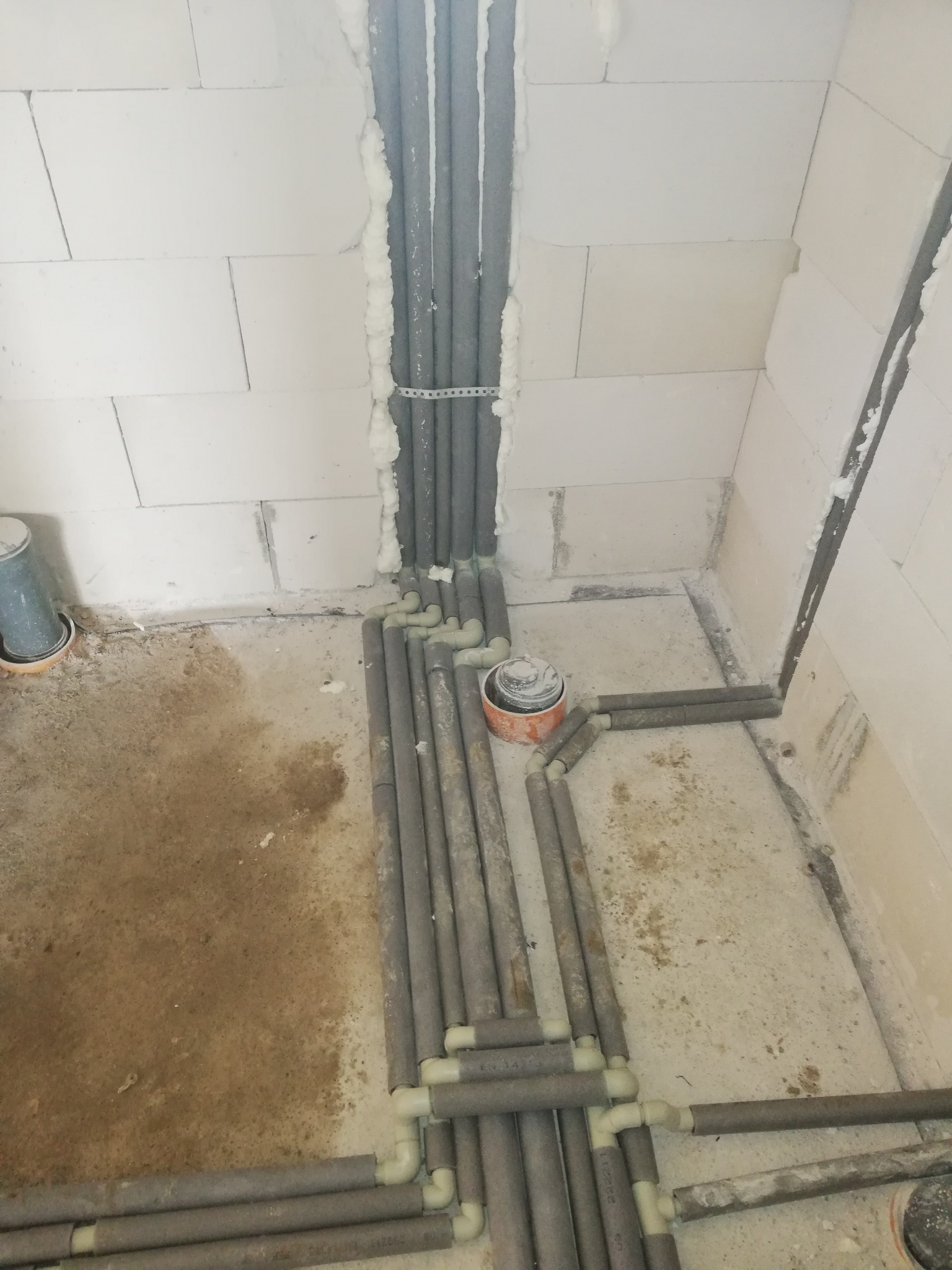 Szare rury instalacji w pianie montażowej, prowadzone wzdłuż ściany z białych bloczków, układane na podłodze w surowym stanie, widoczne złączki.