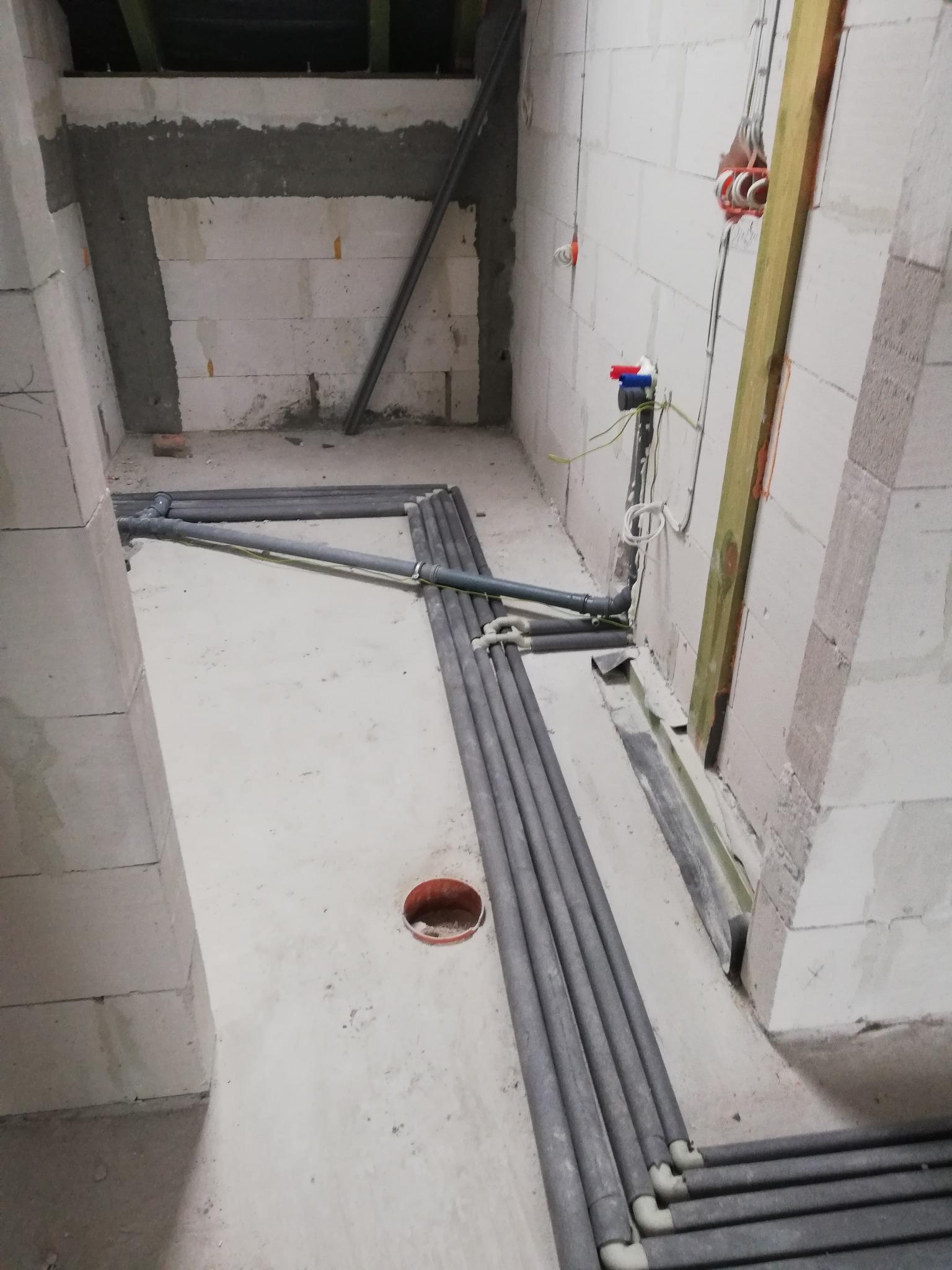Surowy stan: szare rury instalacji w betonie, ściany z białych bloczków, widoczne przewody elektryczne. Wnętrze w trakcie prac instalacyjnych, przed wykończeniem.