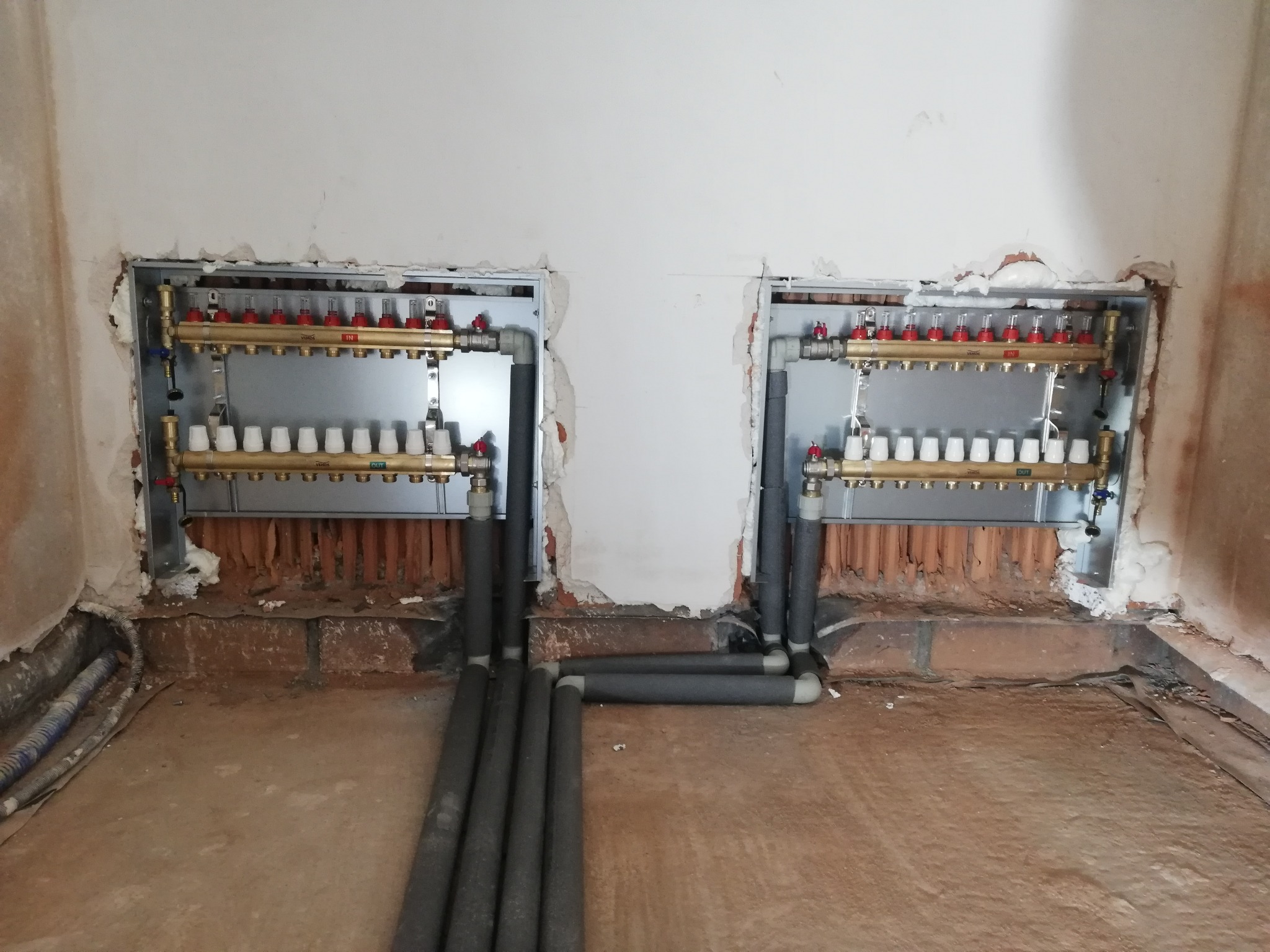 Rozdzielacze ogrzewania podłogowego zamontowane w ścianie, z wyprowadzonymi rurami w szarej izolacji termicznej. Widoczne niedokończone prace wykończeniowe.