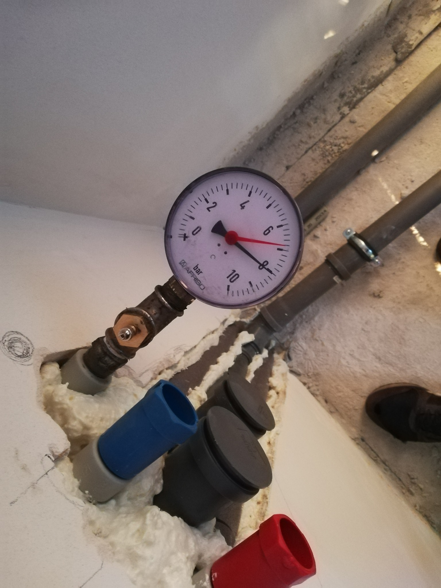 Manometr AFRISO na rurze instalacji hydraulicznej, zabezpieczonej pianką montażową. Widoczne elementy złączek w kolorze niebieskim, czarnym i czerwonym.