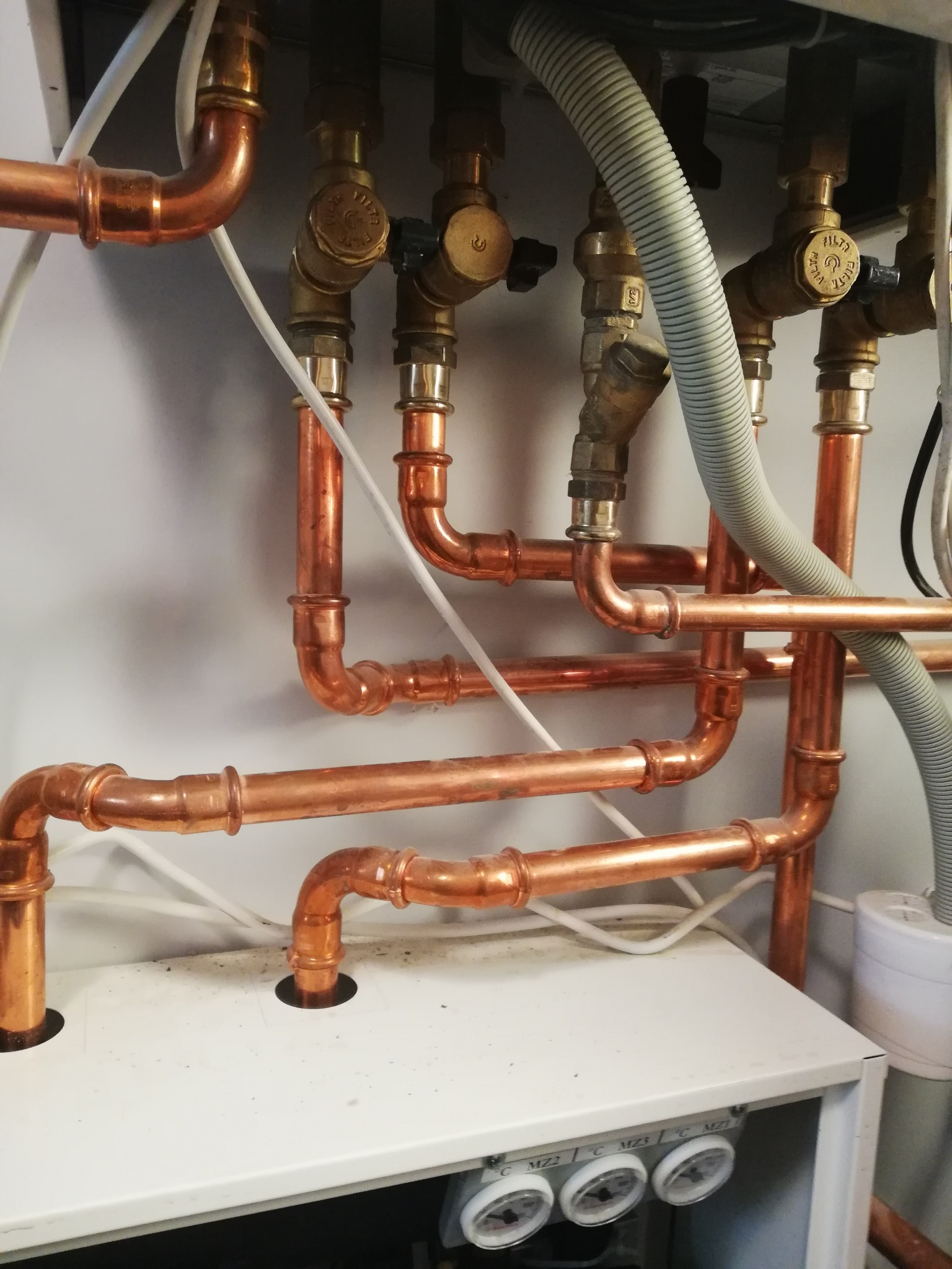 Miedziane rury instalacji grzewczej z zaworami, ułożone na białym panelu. Widoczne złącza lutowane i fragment szarego węża karbowanego.