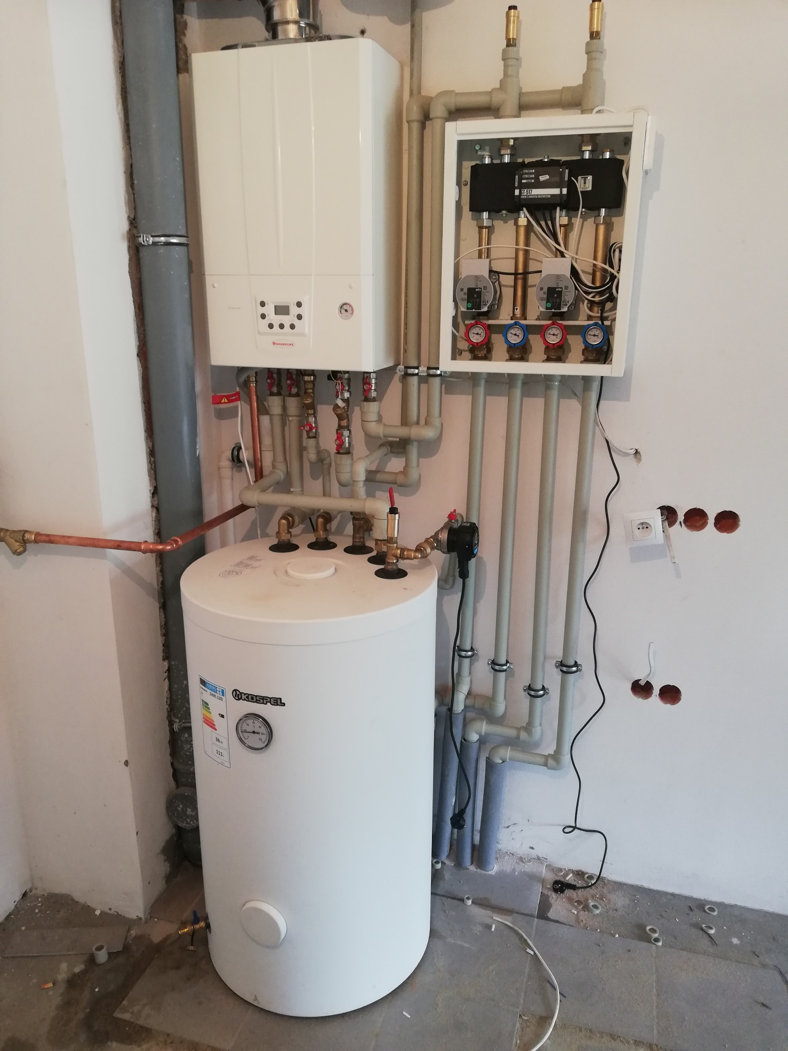 Biały piec gazowy i zasobnik ciepłej wody Kospel w kotłowni, z widoczną instalacją hydrauliczną i rozdzielaczem w białej obudowie. Świeże otwory w ścianie.