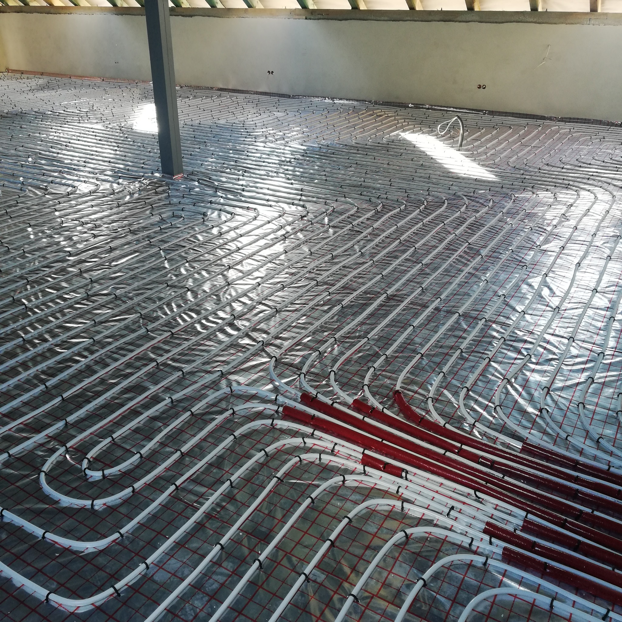 Białe rury ogrzewania podłogowego, częściowo w czerwonej izolacji, ułożone na folii z czerwoną siatką. Widoczny słup podtrzymujący strop i fragment sufitu.