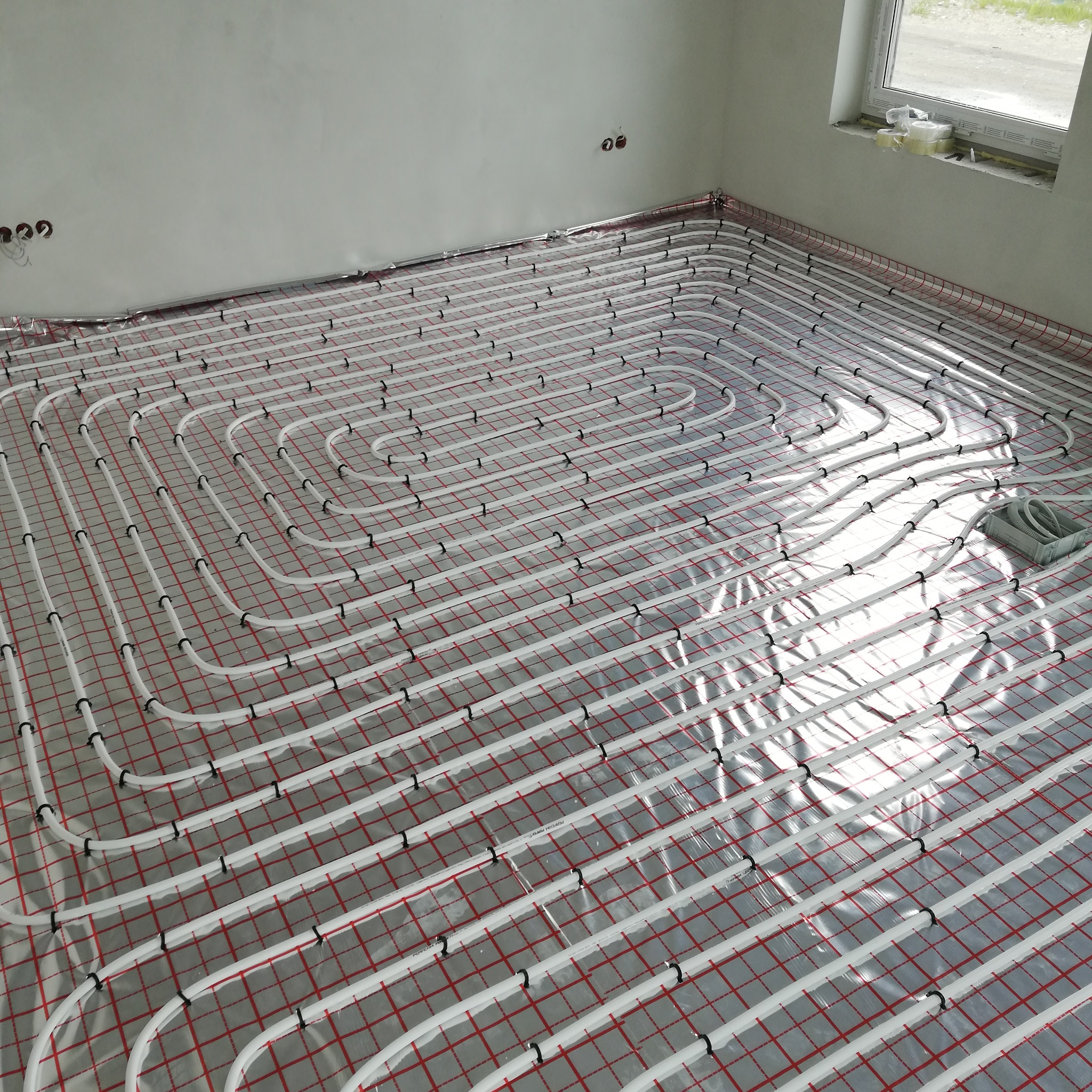 Białe rury ogrzewania podłogowego ułożone na folii z czerwoną siatką w surowym wnętrzu z oknem. Widoczne uchwyty mocujące rury.