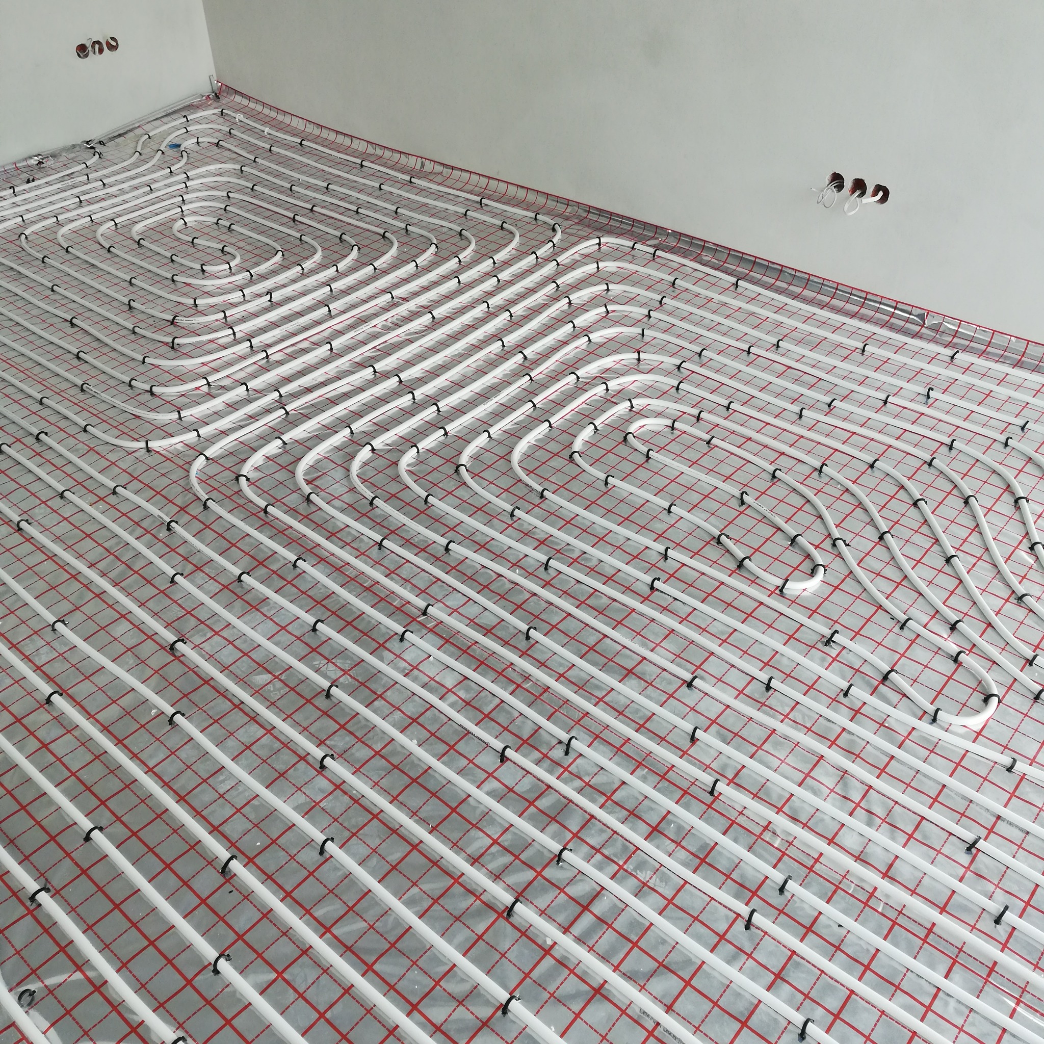 Białe rury ogrzewania podłogowego, ułożone spiralnie na czerwonej siatce, na podłodze w surowym wnętrzu. Widoczne uchwyty mocujące rury.