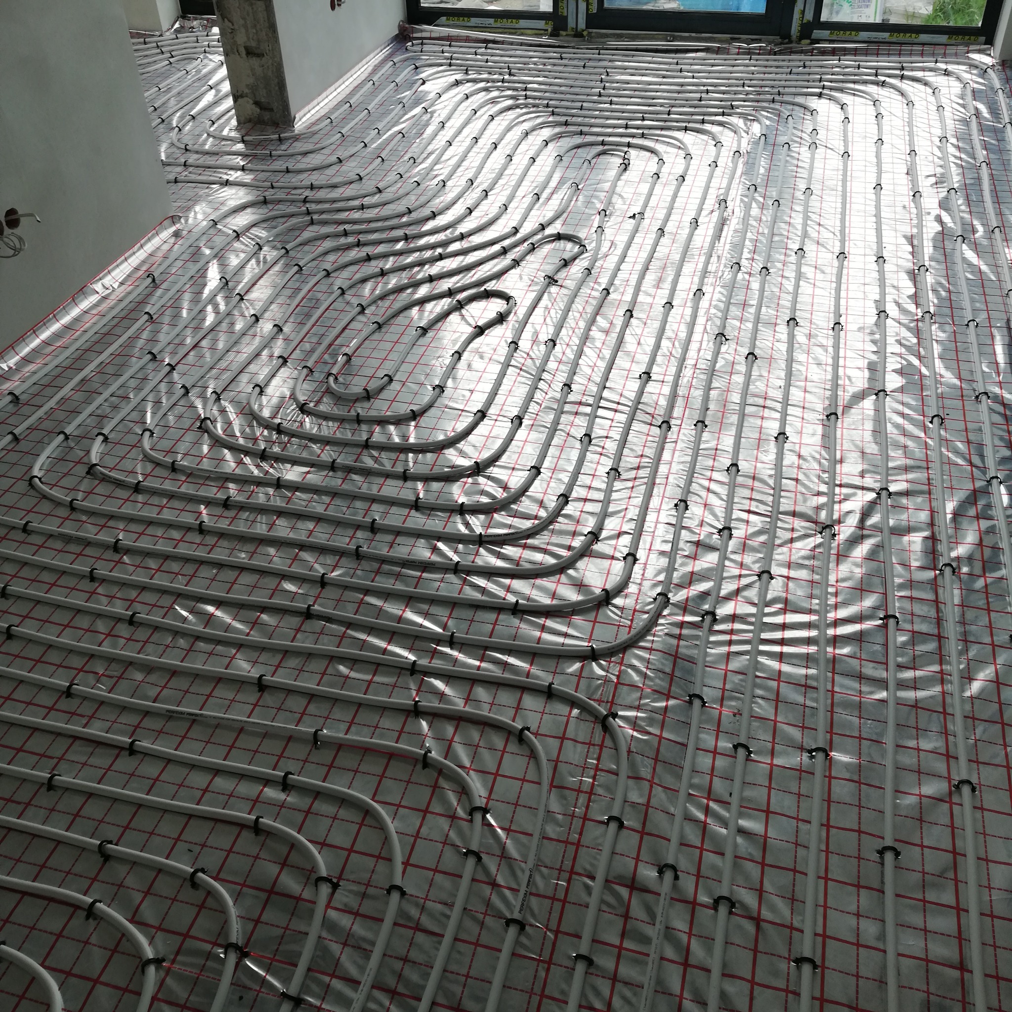 Białe rury ogrzewania podłogowego ułożone na folii z czerwoną siatką, przymocowane czarnymi klipsami, w jasnym pomieszczeniu w trakcie instalacji.