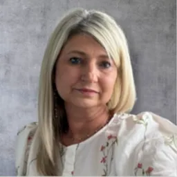 Portret blondwłosej kobiety w kwiecistej bluzce, z delikatnym uśmiechem na tle szarej ściany.