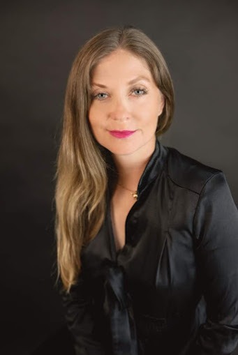 Portret kobiety z długimi blond włosami, w czarnej satynowej bluzce i delikatnym złotym naszyjniku, na ciemnoszarym tle.