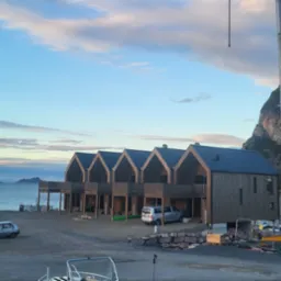 Nowoczesne domki letniskowe z drewnianą elewacją i balkonami, położone nad brzegiem morza, z widocznym krajobrazem górskim w tle i elementami placu budowy.