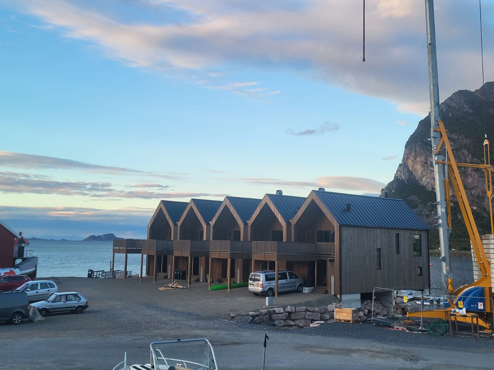 Nowoczesne domki letniskowe z drewnianą elewacją i balkonami, położone nad brzegiem morza, z widocznym krajobrazem górskim w tle i elementami placu budowy.