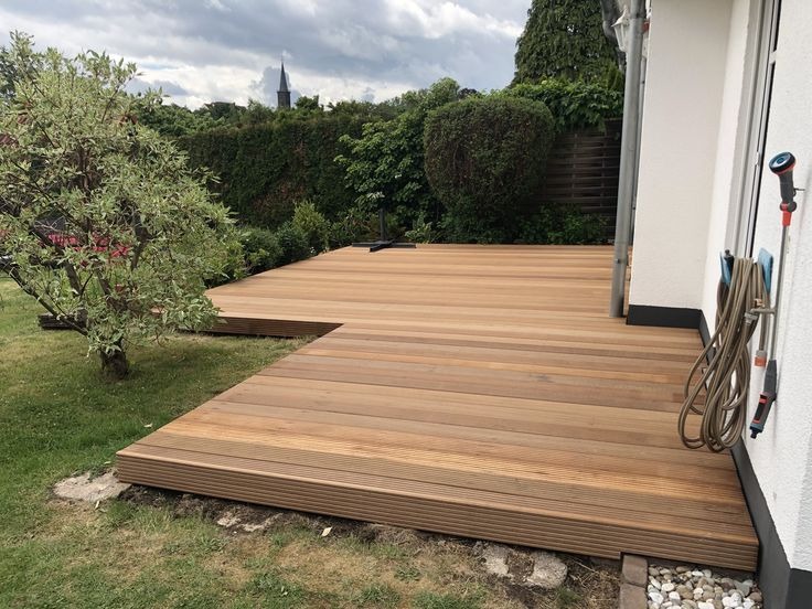 Jasny, drewniany taras z desek ułożonych w regularny wzór, przylegający do białej ściany budynku. W tle zieleń ogrodu i fragment wieży kościoła.