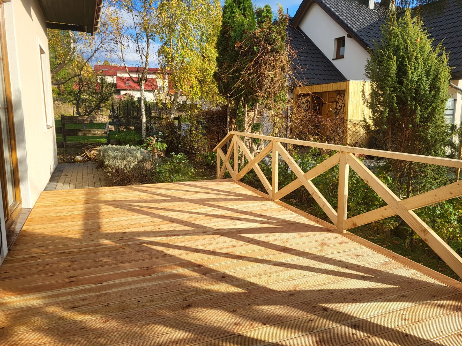 Jasny, drewniany taras z balustradą w geometryczny wzór, oświetlony popołudniowym słońcem. W tle zieleń ogrodu i zabudowa jednorodzinna.