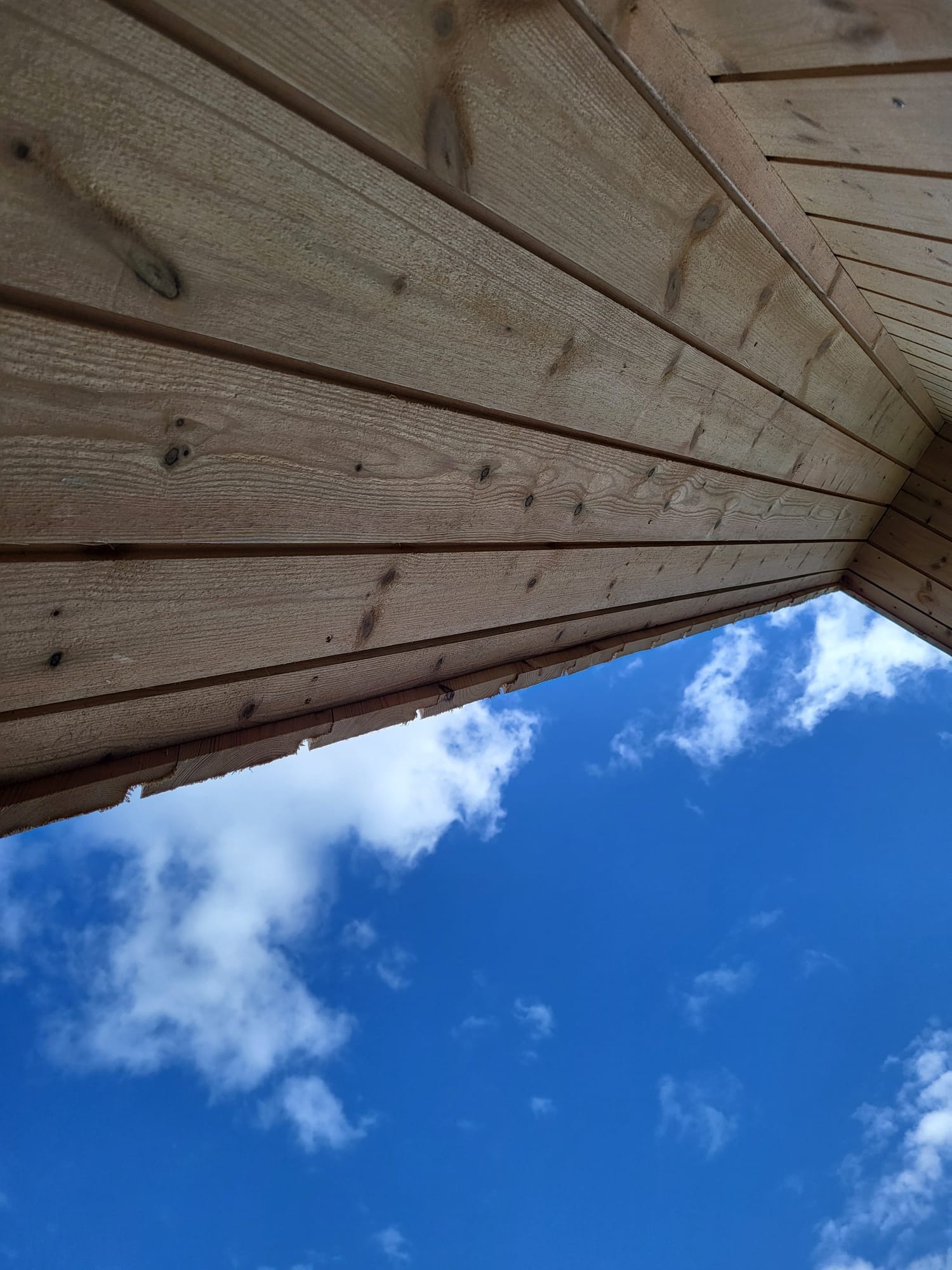 Drewniana konstrukcja od dołu, ukazująca deski z widocznymi słojami i sękami, na tle błękitnego nieba z białymi obłokami. Ujęcie z niskiej perspektywy.
