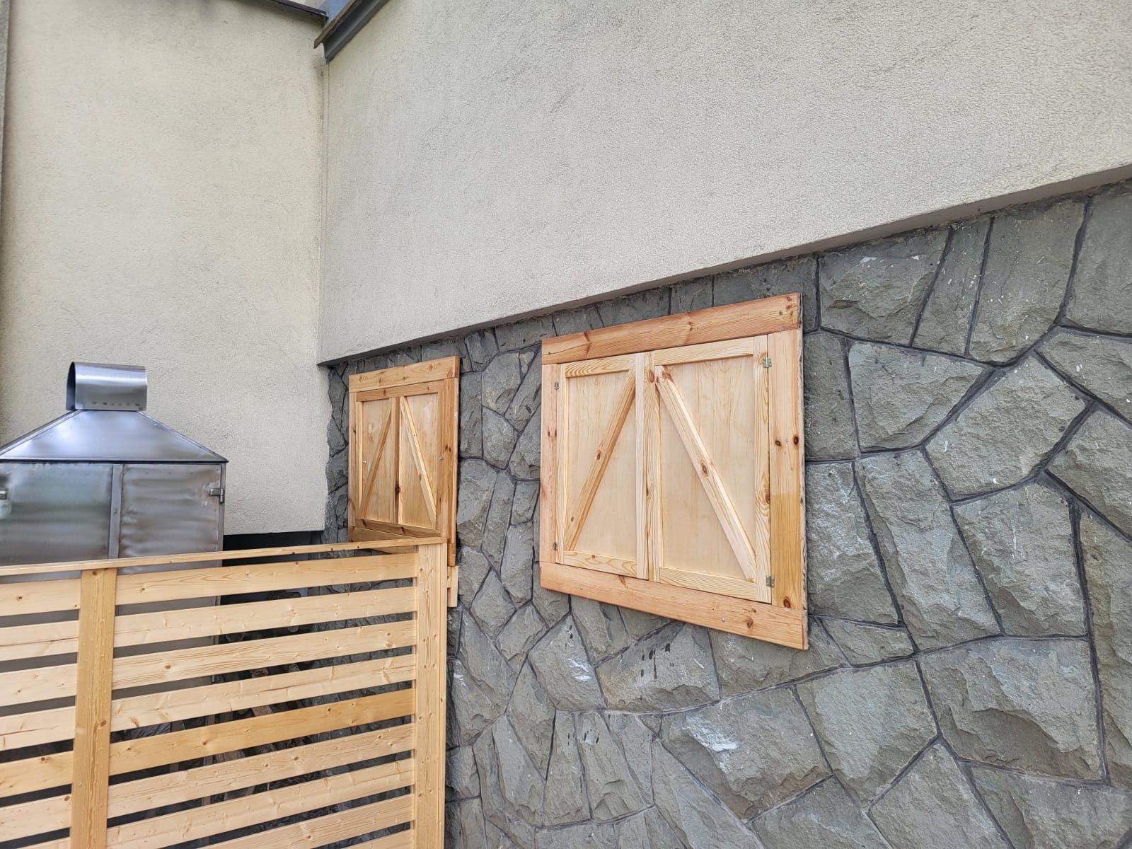 Drewniane okiennice z ukośnymi wzmocnieniami na kamiennej ścianie, obok drewniane ogrodzenie i metalowa konstrukcja. Jasne drewno, szara ściana.