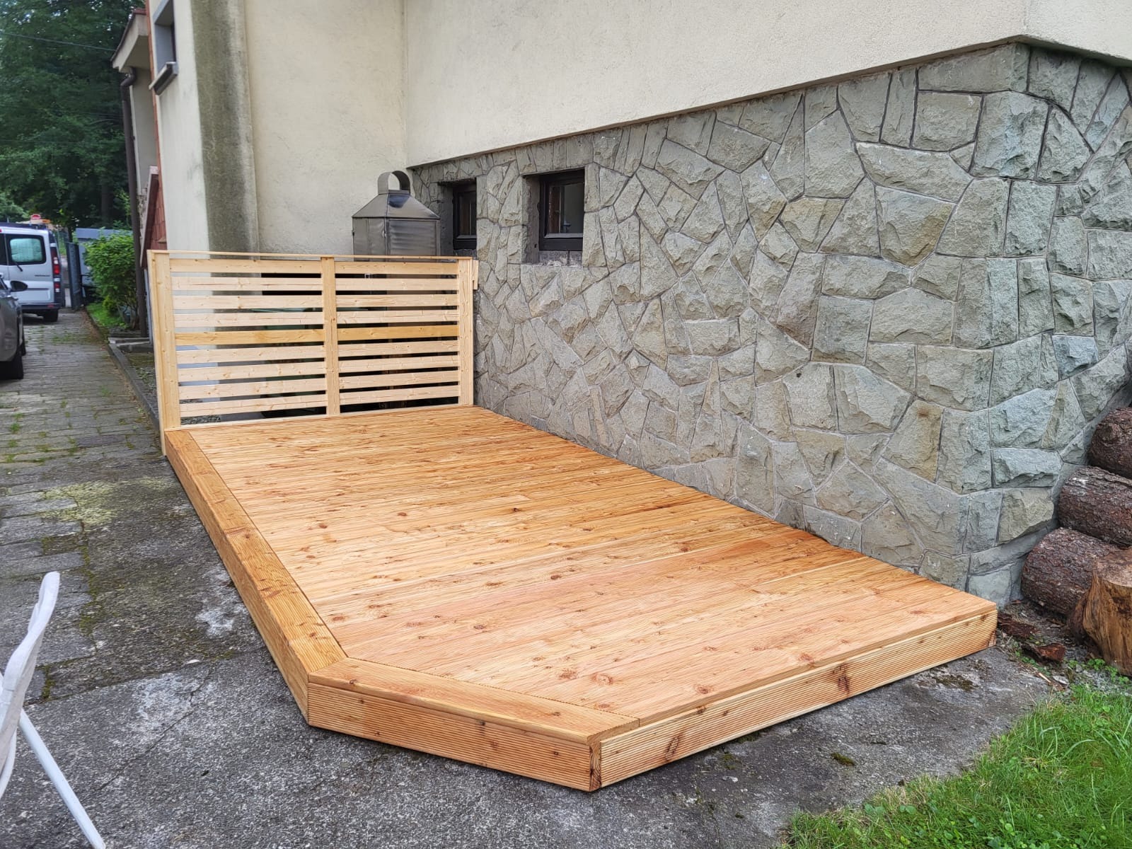 Drewniany taras przydomowy z ażurową ścianką, wykonany z jasnego drewna, na tle kamiennej ściany budynku. Widoczne detale wykończenia i naturalna kolorystyka drewna.