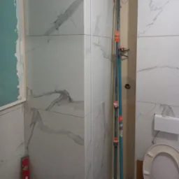 Białe płytki imitujące marmur w trakcie montażu w łazience. Widoczny poziomica, podpórka teleskopowa i fragment toalety. Ściana częściowo przygotowana pod płytki.