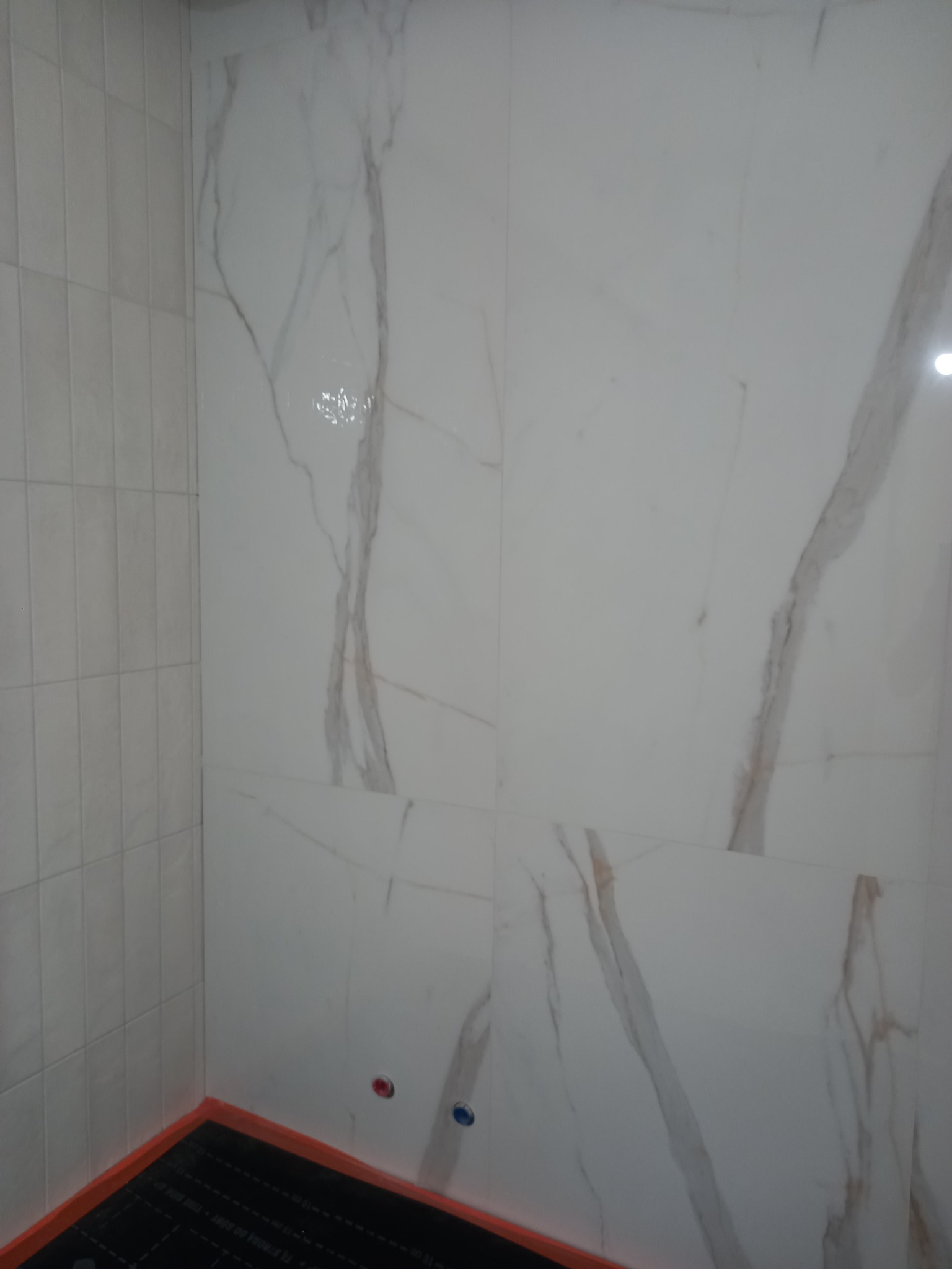 Wykończona ściana łazienki z białych płytek imitujących marmur, z widoczną instalacją ogrzewania podłogowego w narożniku. Minimalistyczny design, ciepłe odcienie.