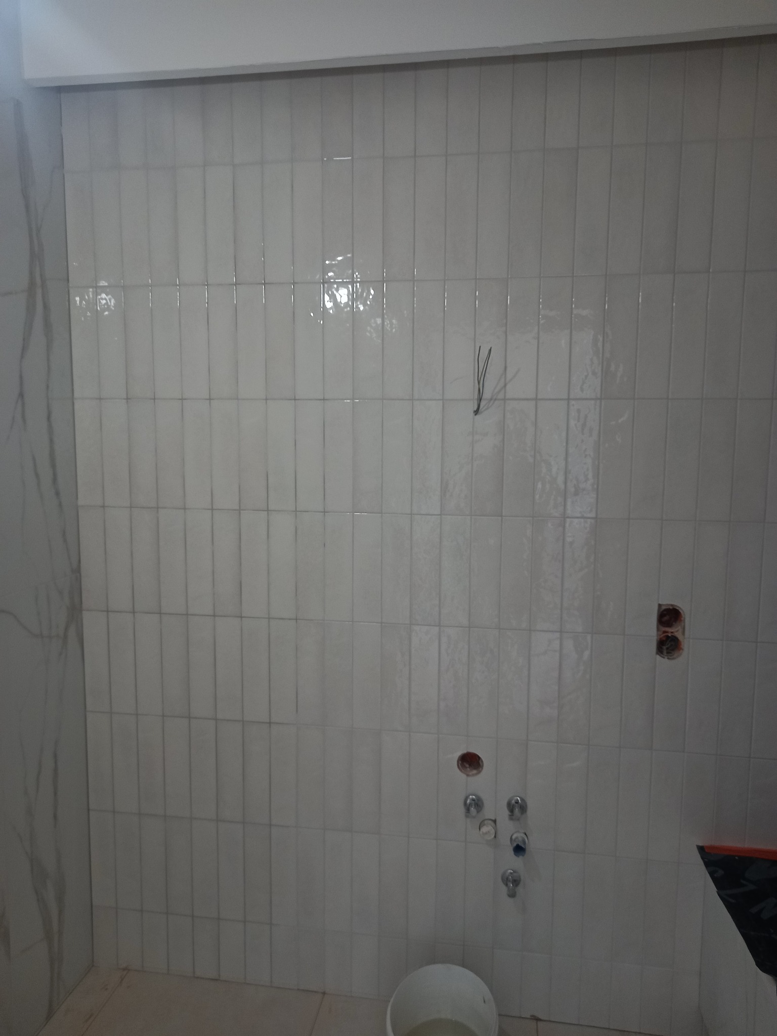 Białe, wąskie płytki ułożone pionowo na ścianie łazienki, widoczne instalacje wodne i elektryczne. Częściowo wykończona ściana z marmurową płytą po lewej.