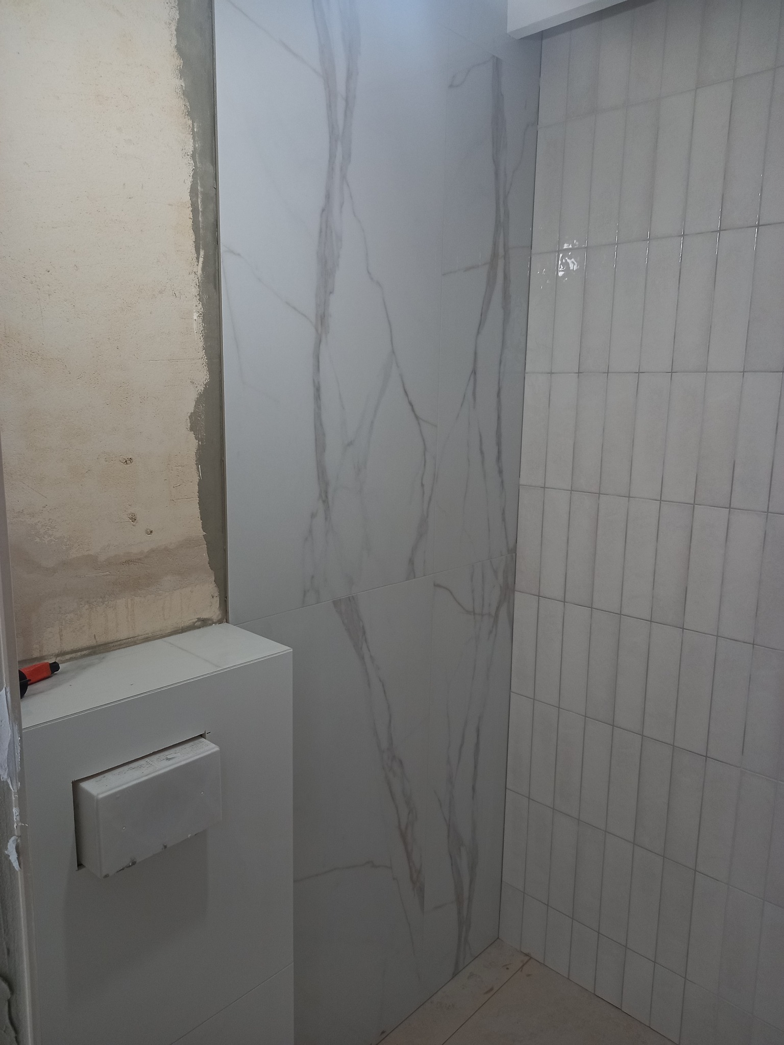 Fragment łazienki w trakcie wykańczania: ściana z imitacją marmuru, pionowe płytki, surowy fragment ściany. Widoczny element instalacji podtynkowej.