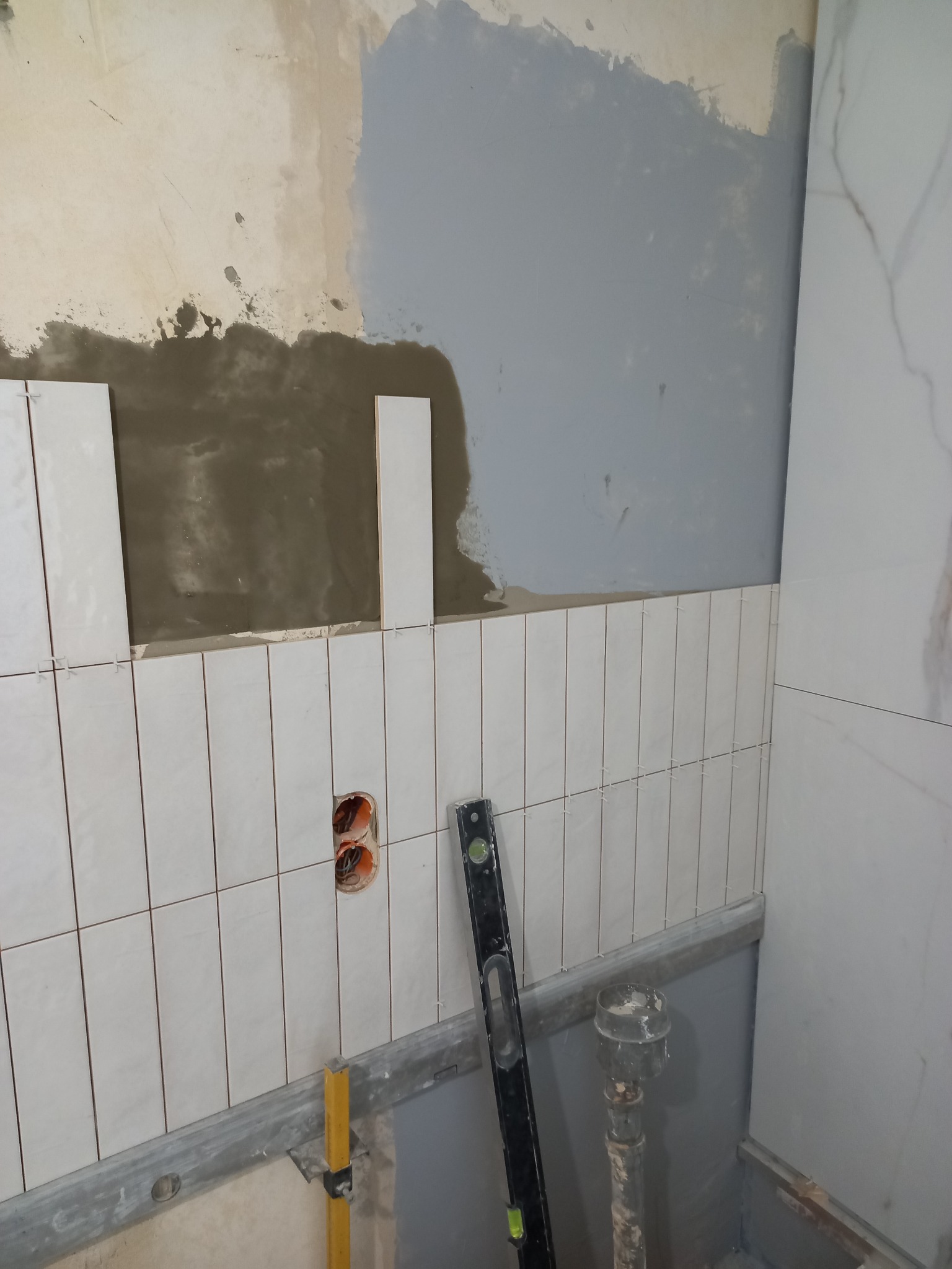 Układanie białych, wąskich płytek na ścianie łazienki. Widoczny poziomica, puszka elektryczna i fragment instalacji wodnej. Prace wykończeniowe w toku.