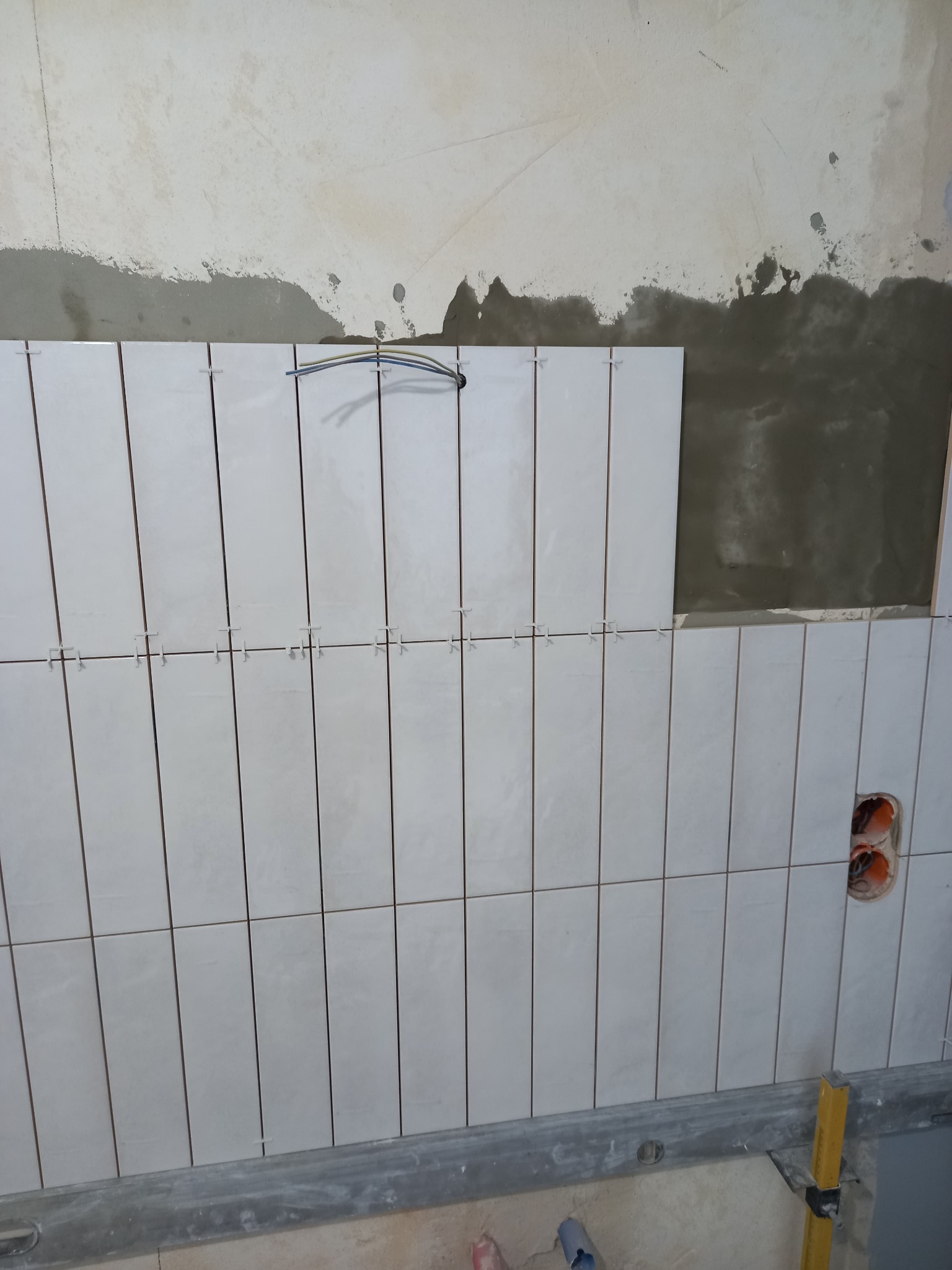 Układanie białych, prostokątnych płytek na ścianie z użyciem krzyżyków dystansowych. Widoczne przewody elektryczne i poziomica.